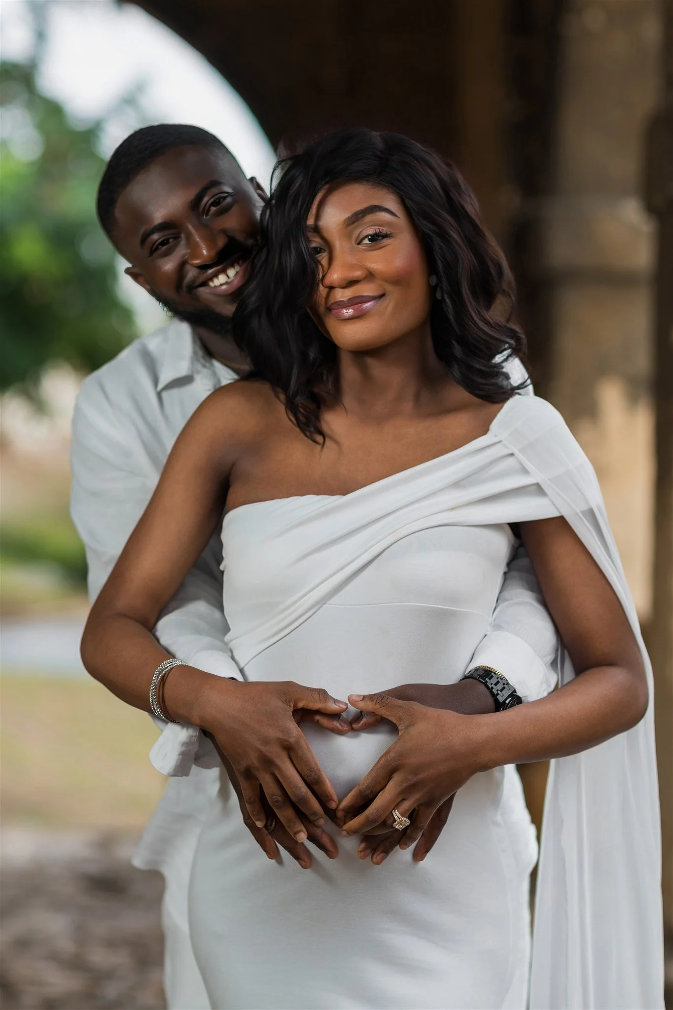 A smiling couple in white stands lovingly under an archway. The man embraces the woman from behind, both forming a heart over her pregnant belly. Serene joy.