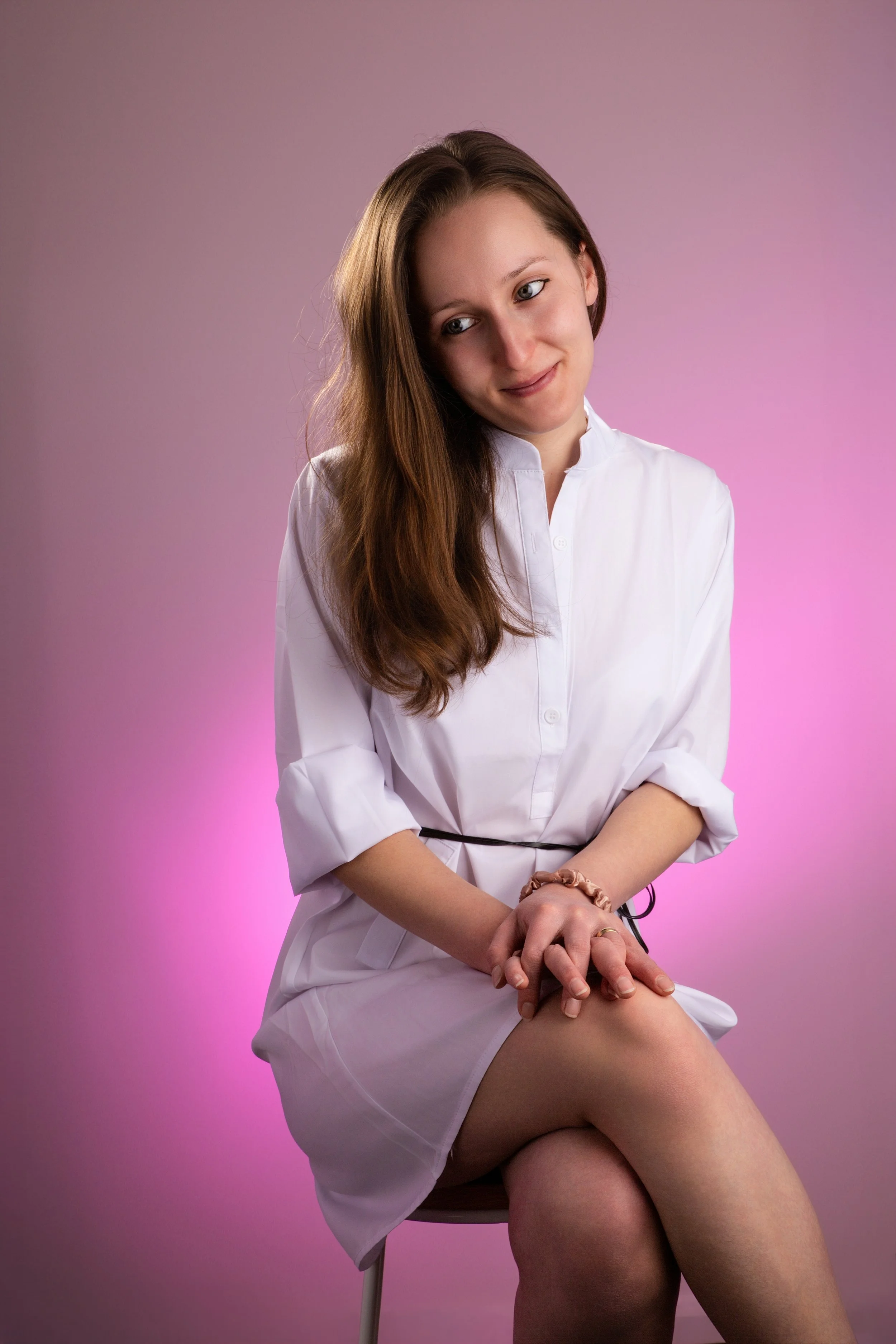 Indoor portrait of a young woman with brown hair in a white dress against a pink background 
