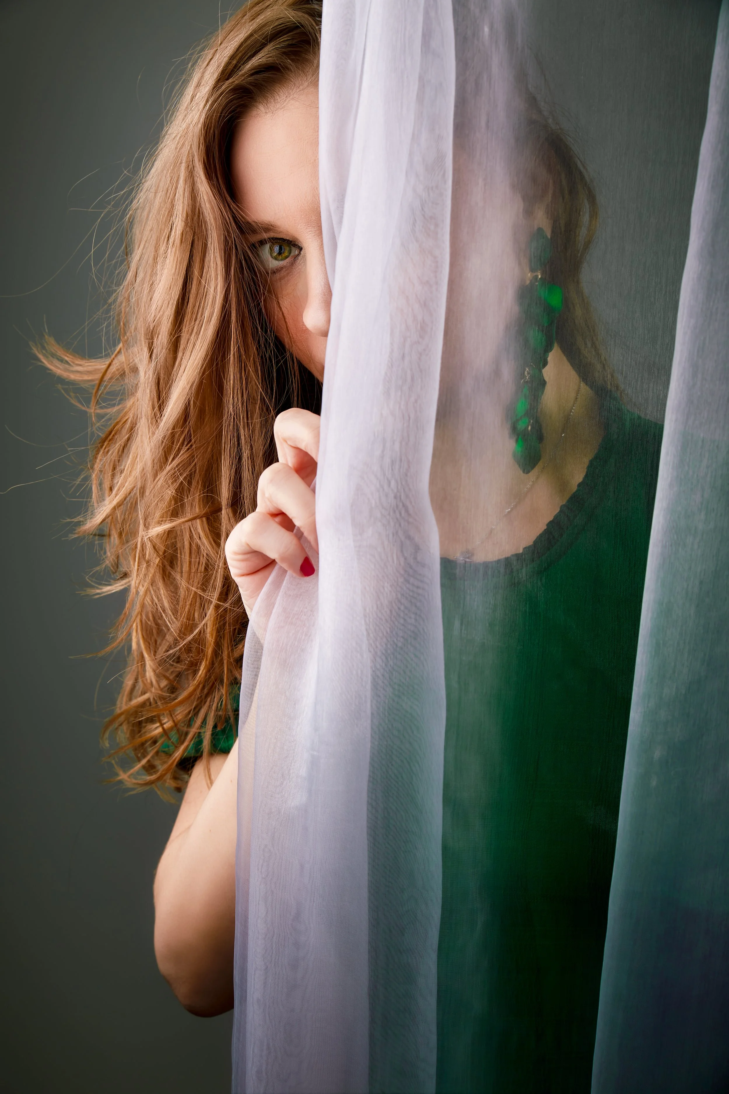 Indoor portrait of a light brown hair woman hiding behind a mesh curtain wearing green dress and green earrings 