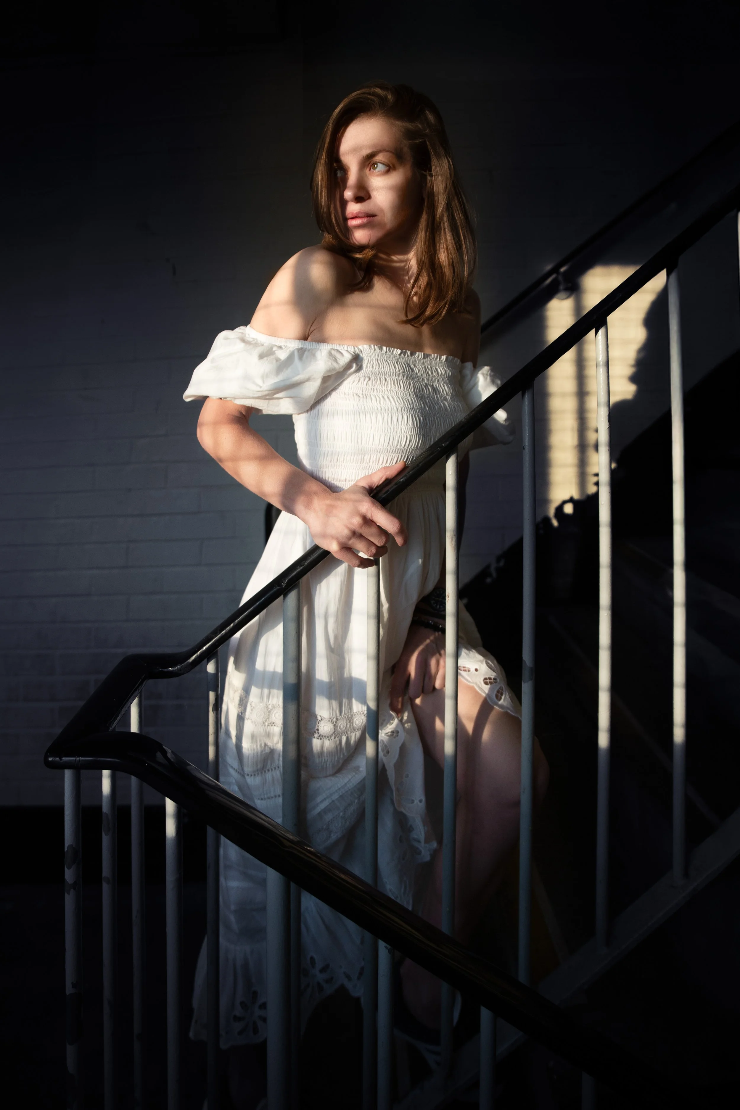 Moody portrait of brown hair young woman wearing white summer dress on a staircase in an industrial setup