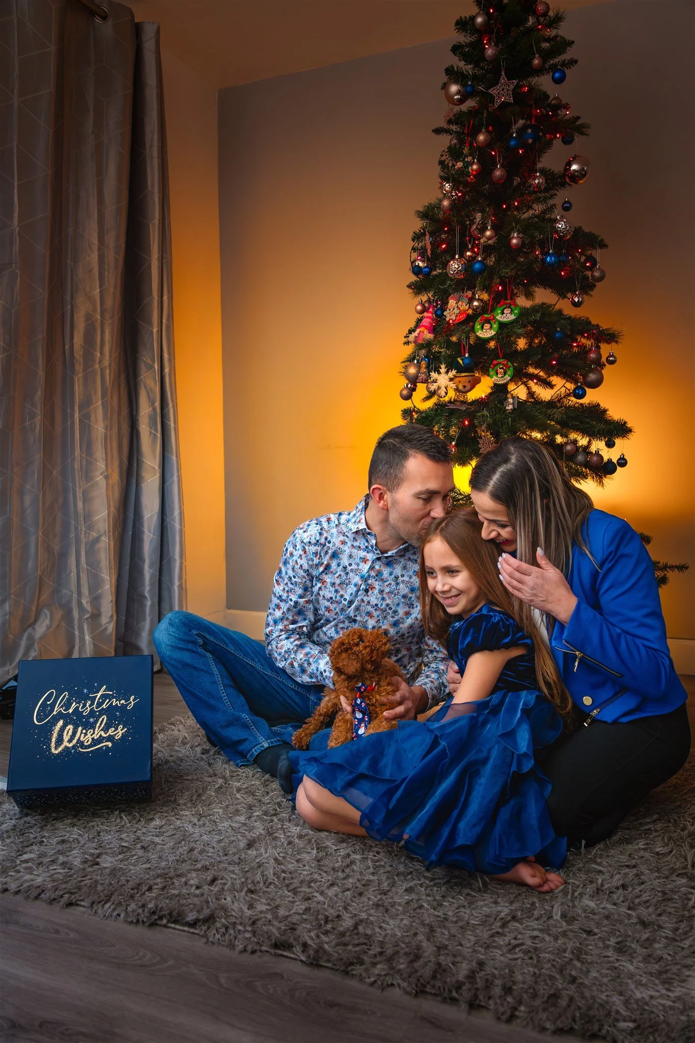 A family sits on a rug by a Christmas tree, sharing a joyful moment. The child, holding a small brown dog, is hugged by the parents. A "Christmas Wishes" box is nearby.
