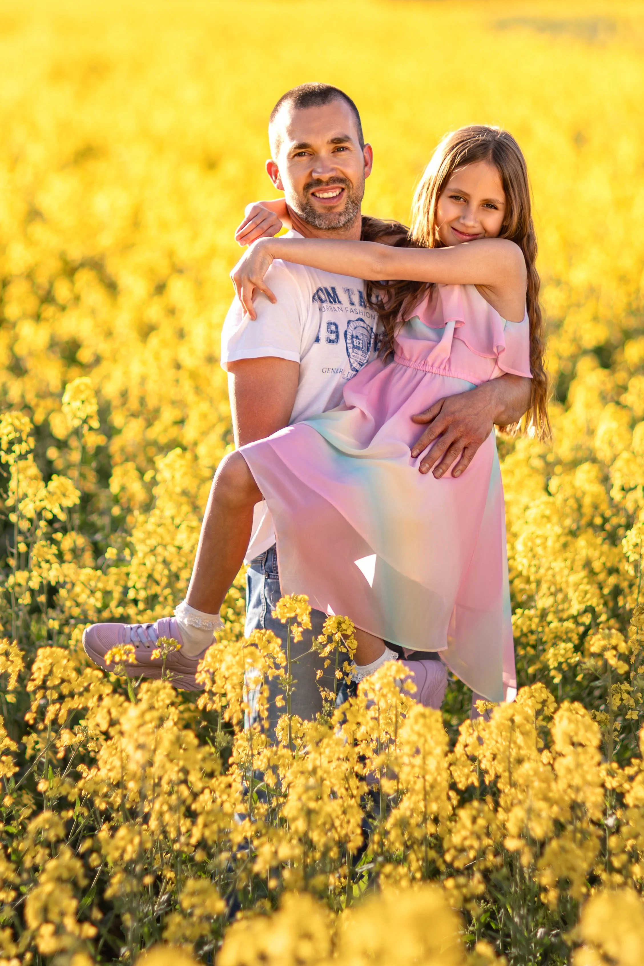 A smiling father holding his daughter in a blooming yellow rapeseed field during golden hour, surrounded by warm sunlight and soft spring colors.