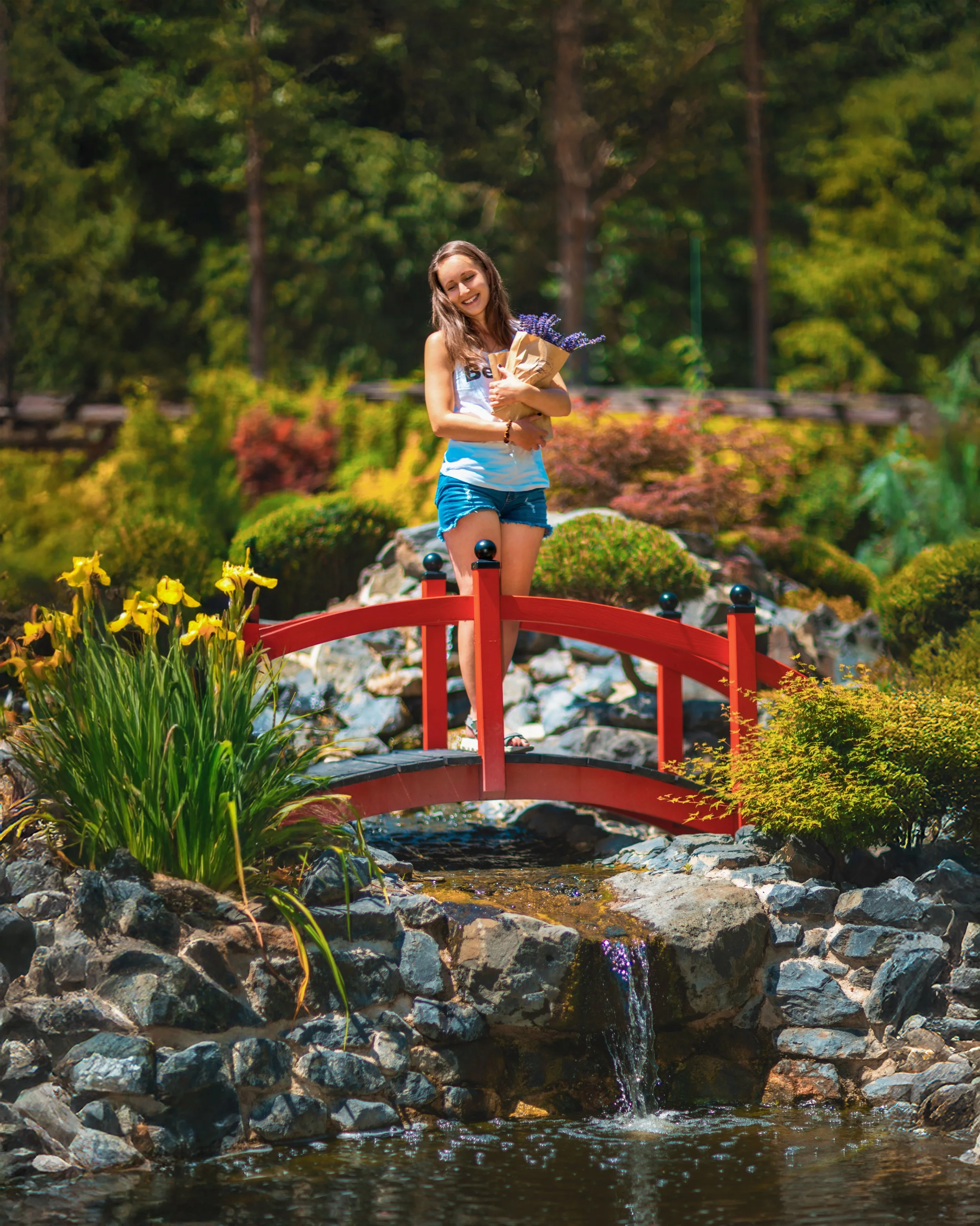 Summery portrait of a young woman standing on a red bridge over a small creek surrounded by rocks and yellow flowers