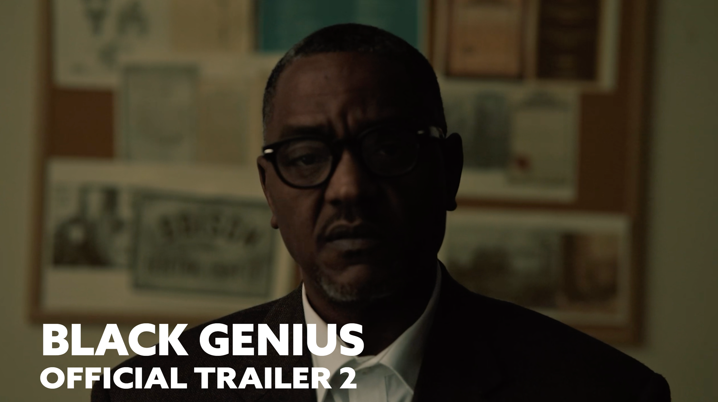 Man with glasses in a dimly lit room, background with framed pictures or posters, text overlay reads 'BLACK GENIUS OFFICIAL TRAILER 2'.