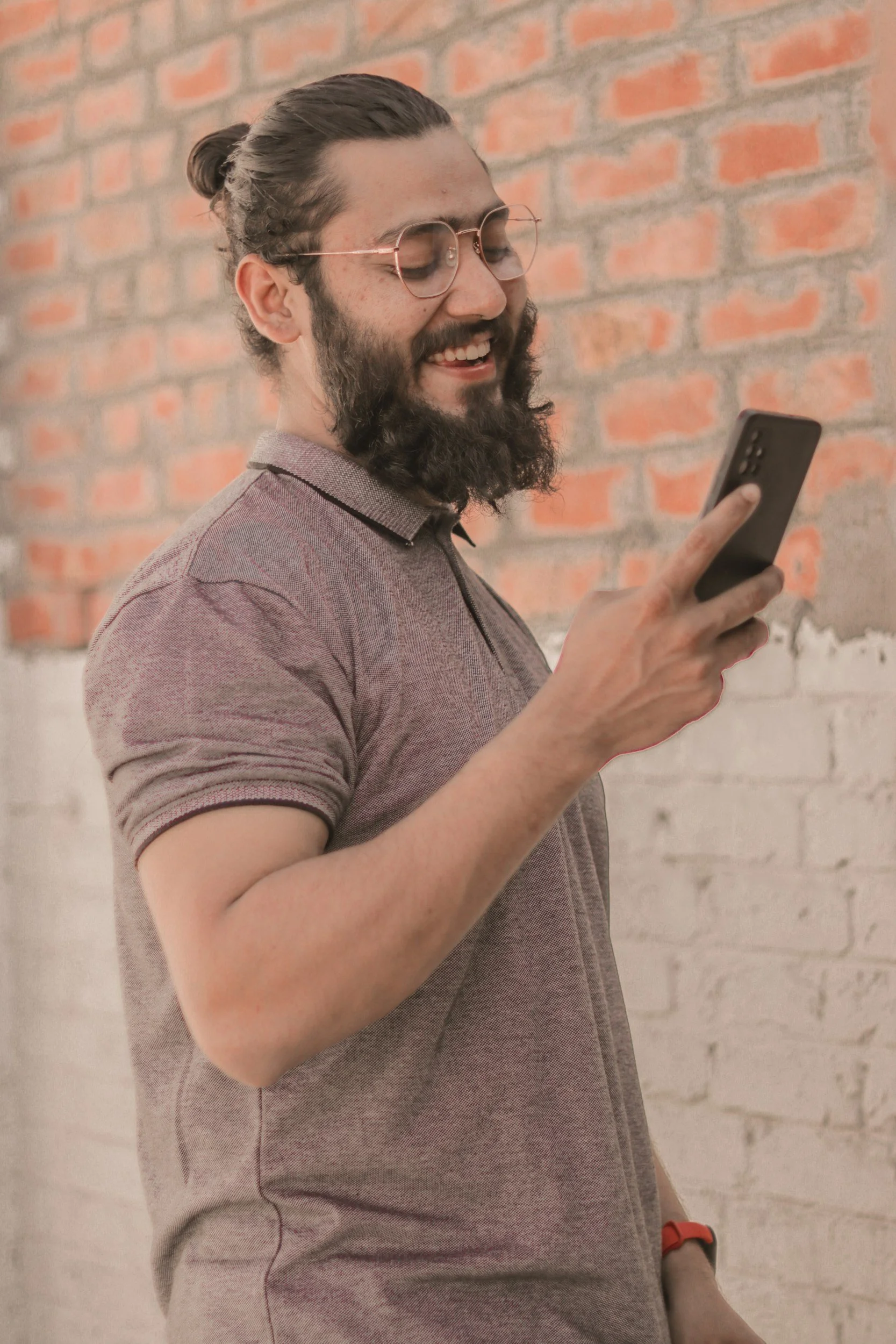 A man with long hair in a bun, glasses, and a beard, smiling while looking at his smartphone, standing against a brick wall.