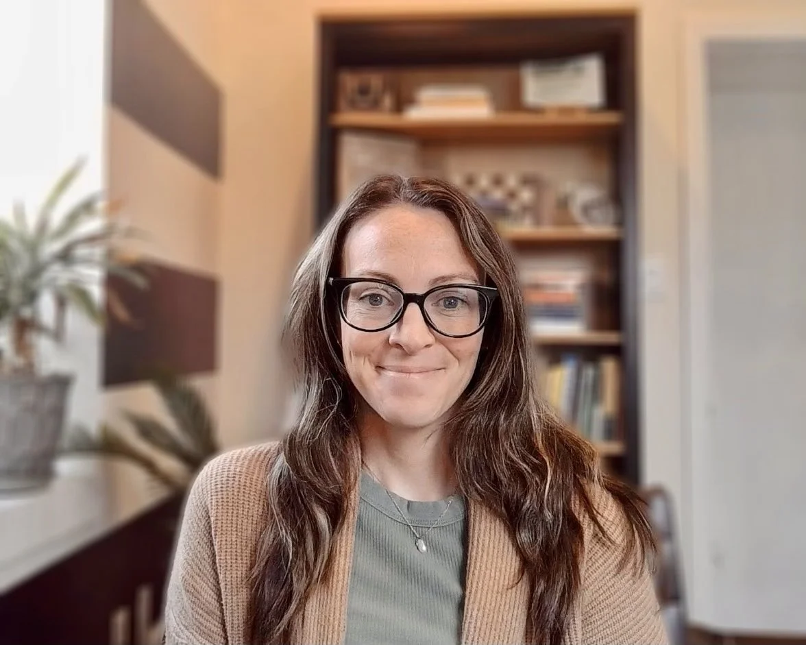 A woman with long brown hair, glasses, and a beige cardigan smiling at the camera in a room with a bookshelf and potted plant in the background.
