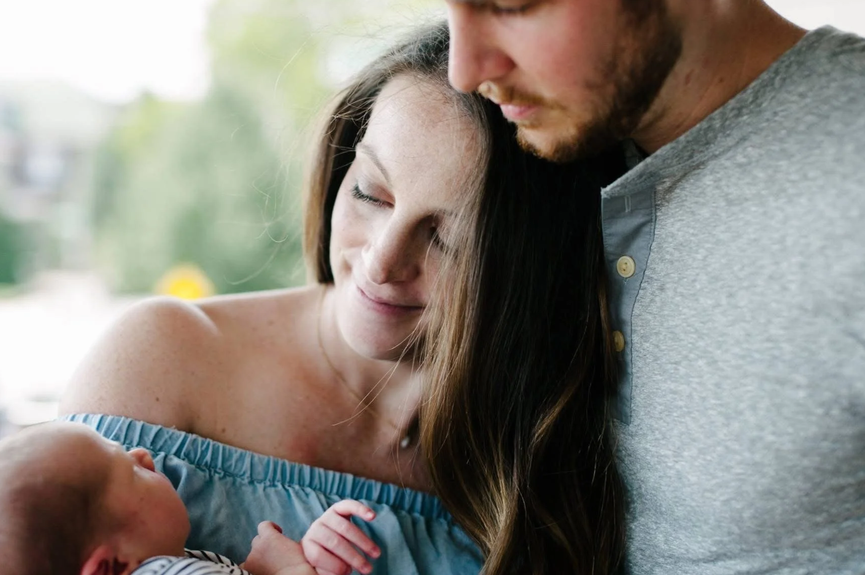 A woman with long brown hair, and a man with facial hair, are seen closely together holding a newborn baby, with the woman gently smiling and the man gently smiling. The background is blurred with greenery.