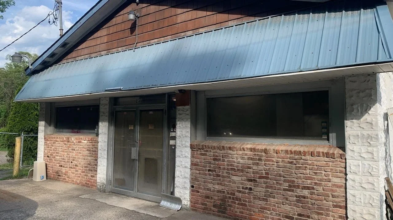 Exterior of a building with brick and stone walls, large windows, and a metal awning over the entrance, with a small concrete area in front.