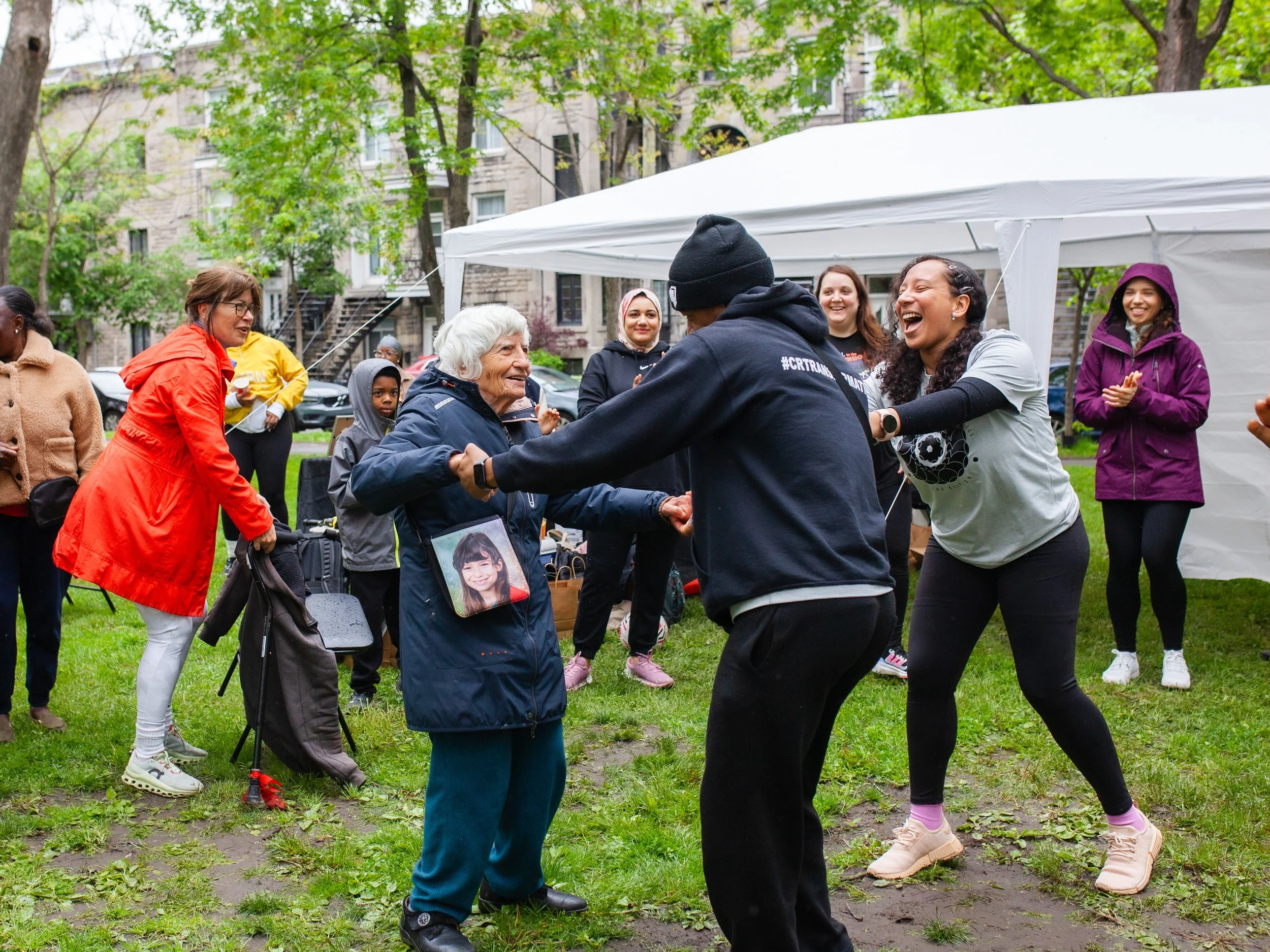 Groupe de personnes qui dansent et rient lors d'une fête en extérieur sous une tente blanche, avec des arbres et des bâtiments en arrière-plan.