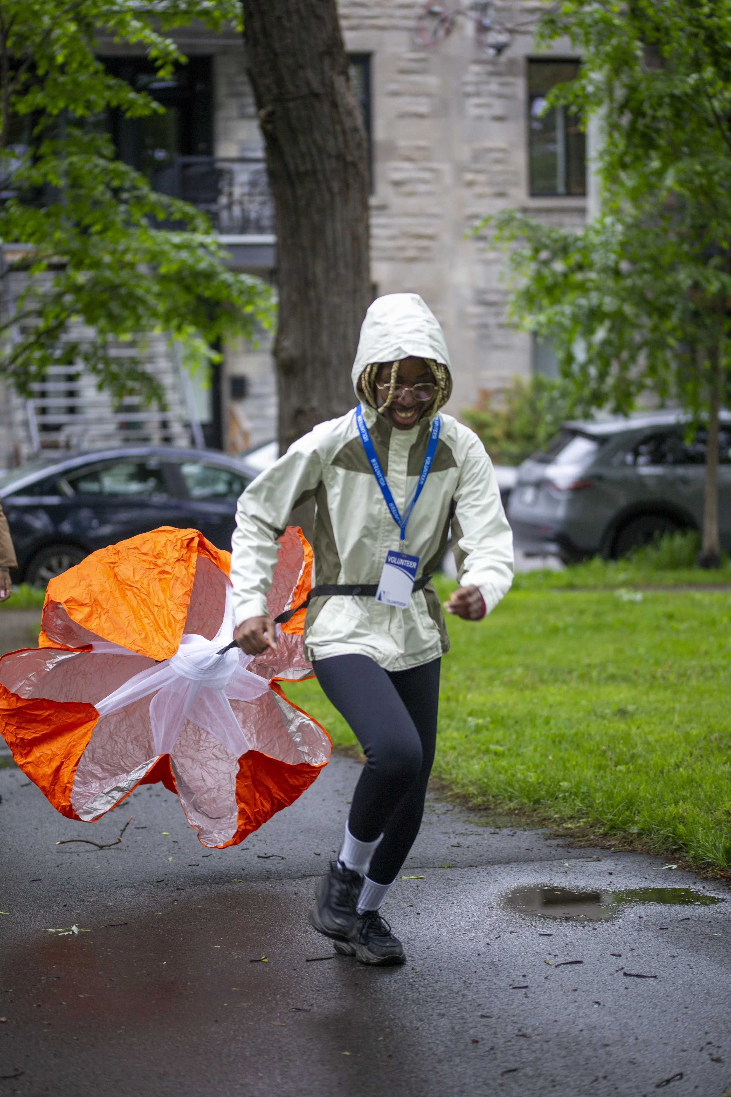 Une personne souriante en veste blanche, portant un badge de bénévole, court dans un parc après une pluie, tenant un cerf-volant orange et blanc.