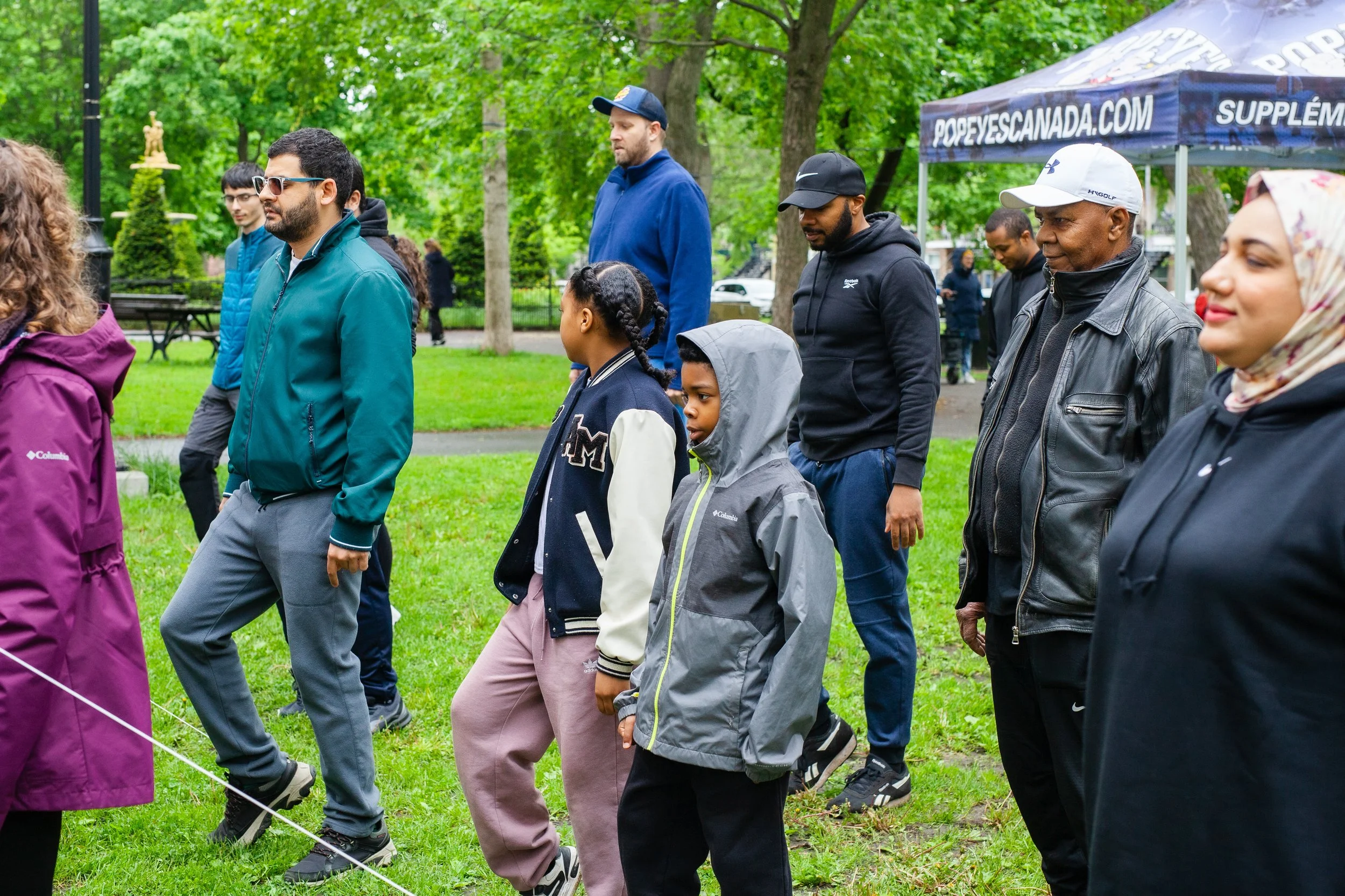 Groupe de personnes de différentes ethnies marchant dans un parc verdoyant sous des arbres, portant des vêtements décontractés et des casquettes, avec une tente en toile de fond portant le site web popeyescanada.com.