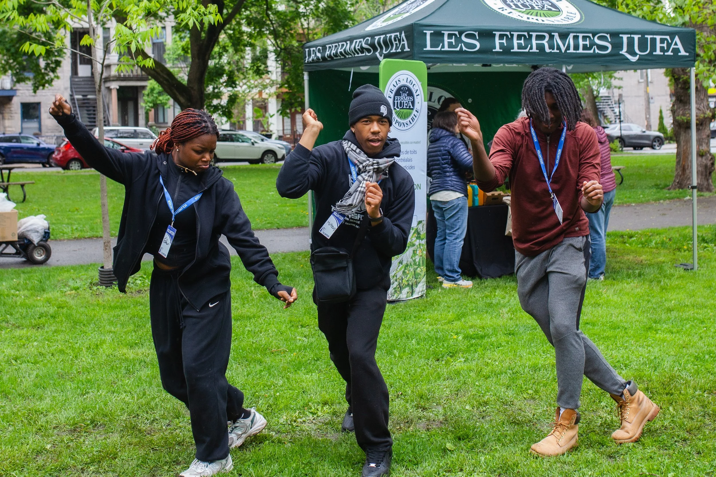 Trois personnes dansent sur l'herbe lors d'un événement en plein air, avec un stand sous une tente verte en arrière-plan.
