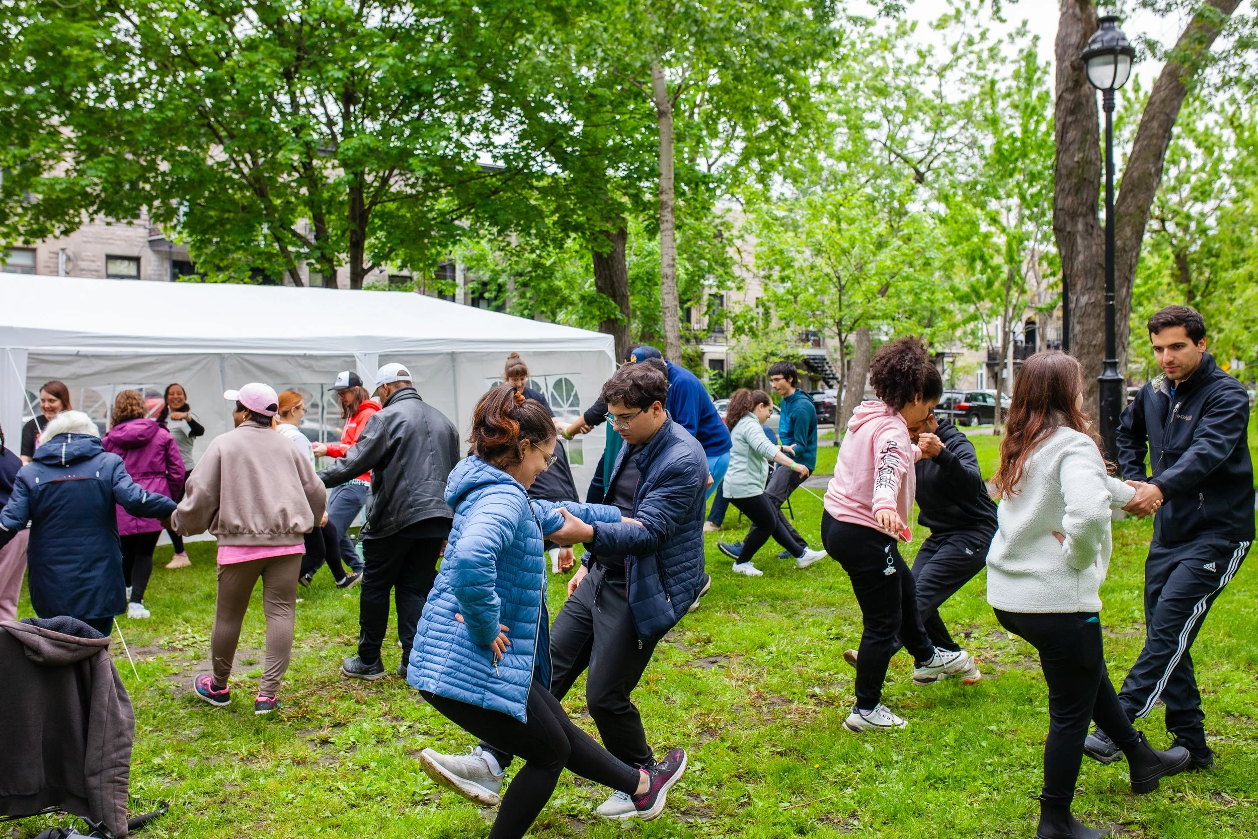 Des personnes dans un parc participent à une fête ou un événement sous une tente blanche, avec des arbres en arrière-plan, en train de danser et de se faire des câlins.