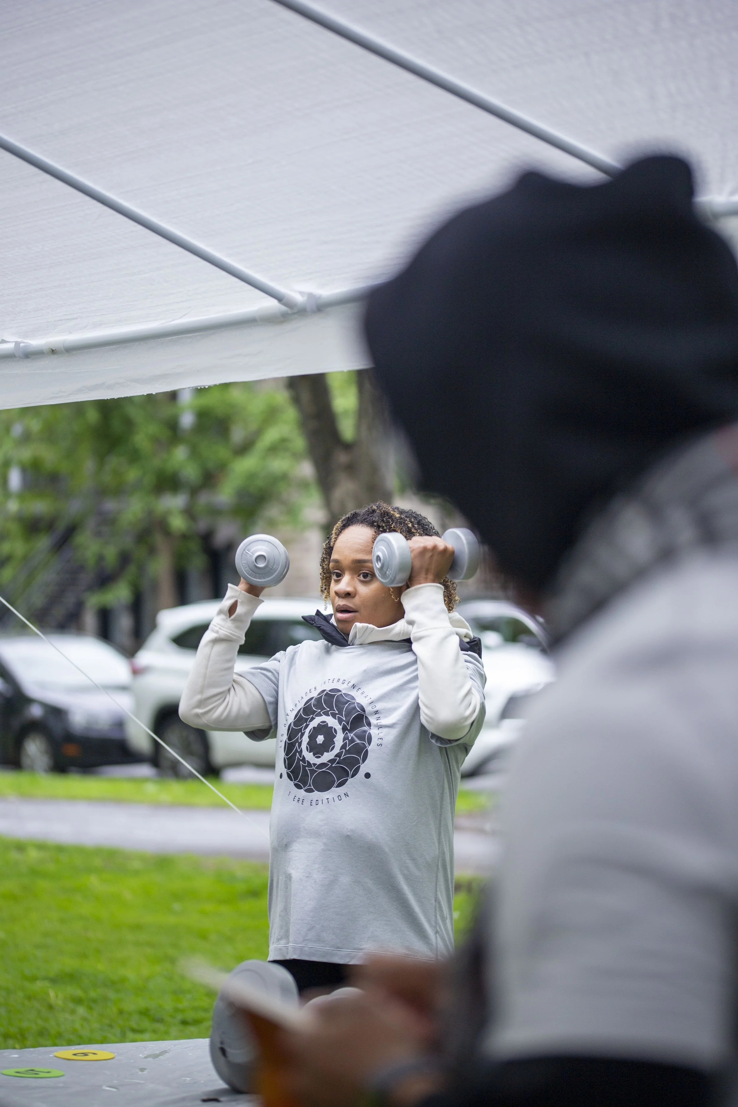 Une femme qui fait des exercices avec des haltères en extérieur, sous une tente, avec un homme équipé d'une montre ou d'un appareil électronique devant elle.