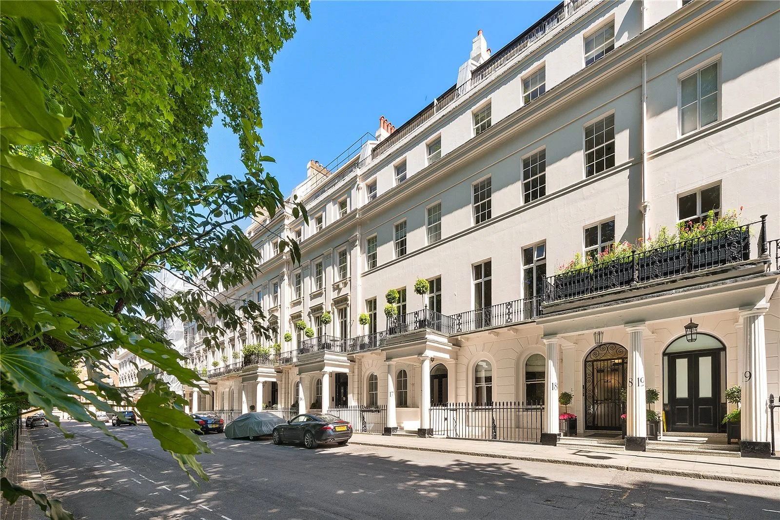 A row of white townhouse buildings with black accents, iron balconies, and potted plants, viewed from a tree-lined street with parked cars on a sunny day.