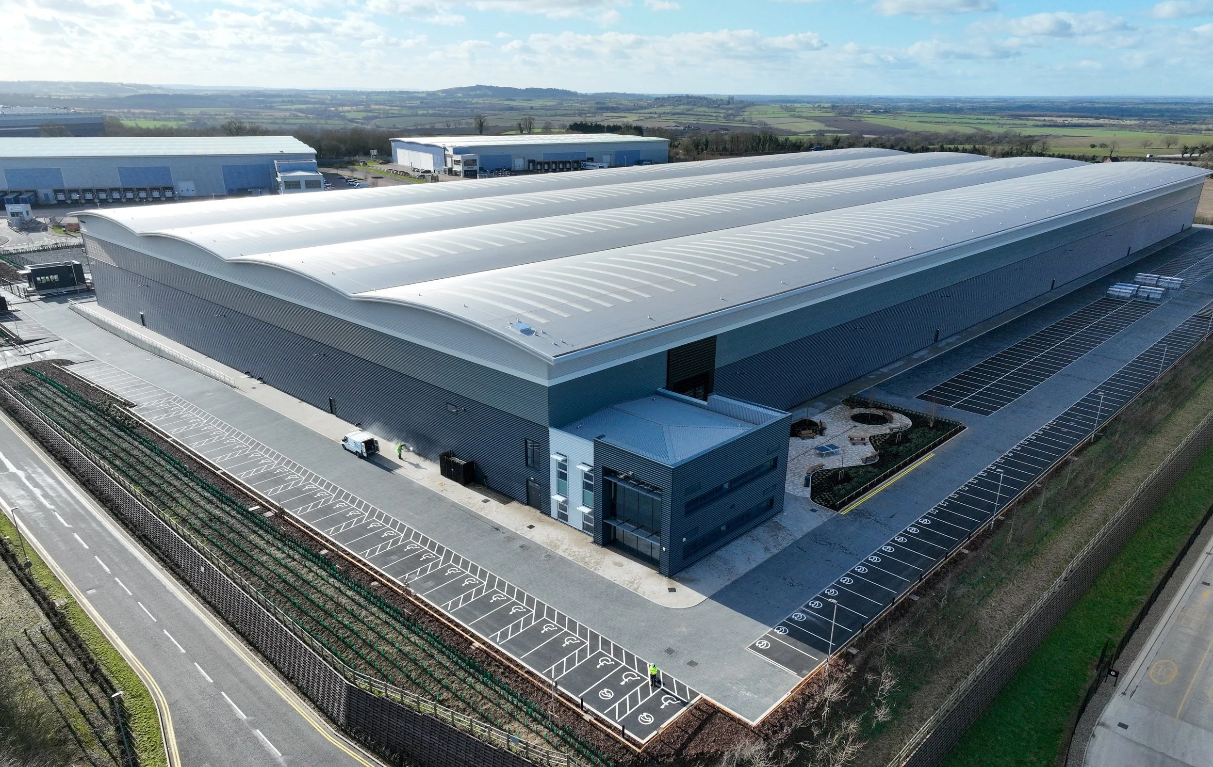 Large industrial warehouse with a shiny white roof, surrounded by parking lots and roads, with a scenic countryside in the background.