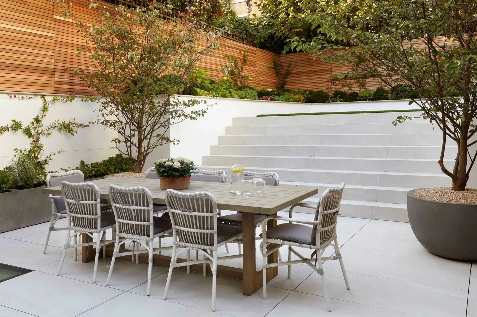 Outdoor patio with a wooden dining table surrounded by seven chairs, potted plants, trees in large planters, stone steps leading up to a garden with a wooden fence and various plants.
