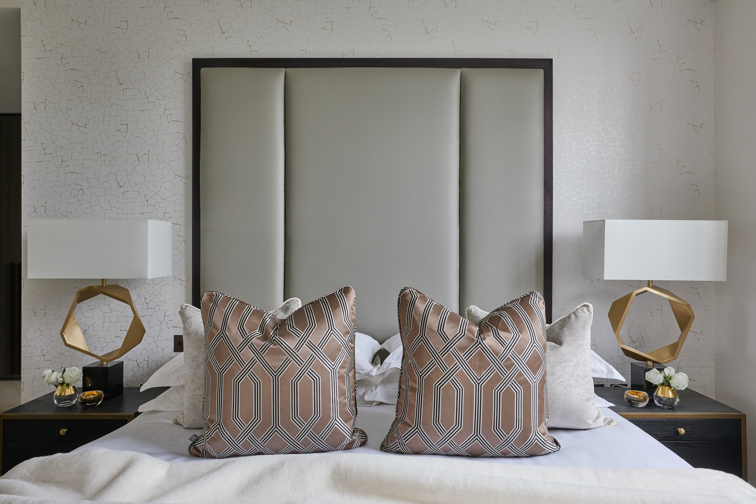 A modern bedroom with a bed that has a tall, padded headboard and four decorative pillows with geometric patterns. There are matching bedside tables with hexagonal lamps and small vases with flowers. The wall behind the bed has a textured, crackle-li