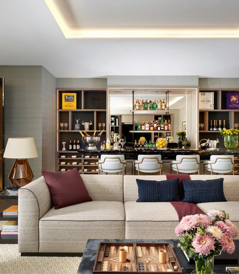 Modern living room with beige sofa, pink and navy throw pillows, floral arrangement, and black coffee table with a backgammon game, facing a bar with wine bottles, glasses, and liquor bottles, flanked by built-in shelves with decorative items and boo