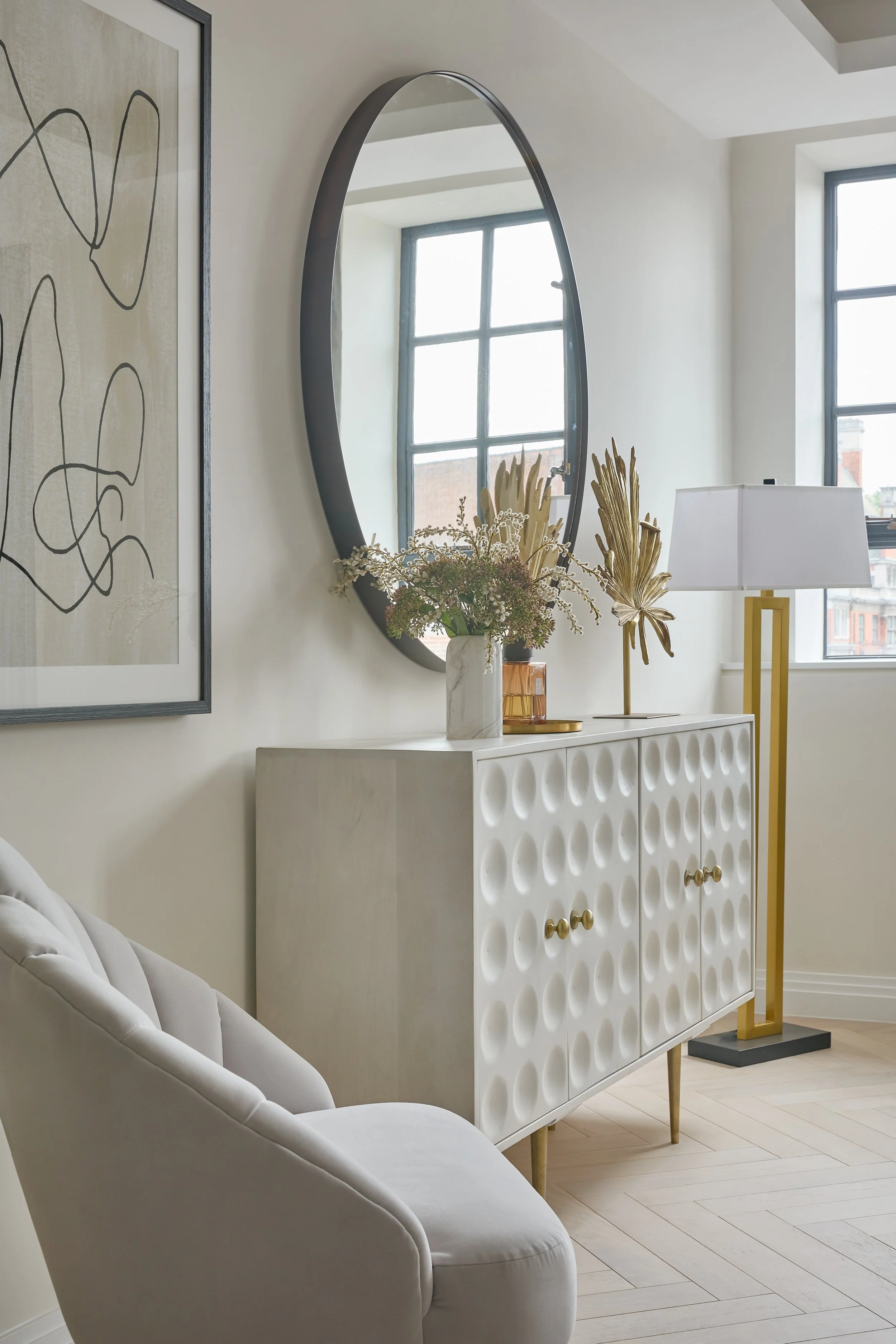 Interior of a modern living space with a white sideboard, a round mirror, a floor lamp with a white shade, and decorative plants, with large windows letting in natural light.