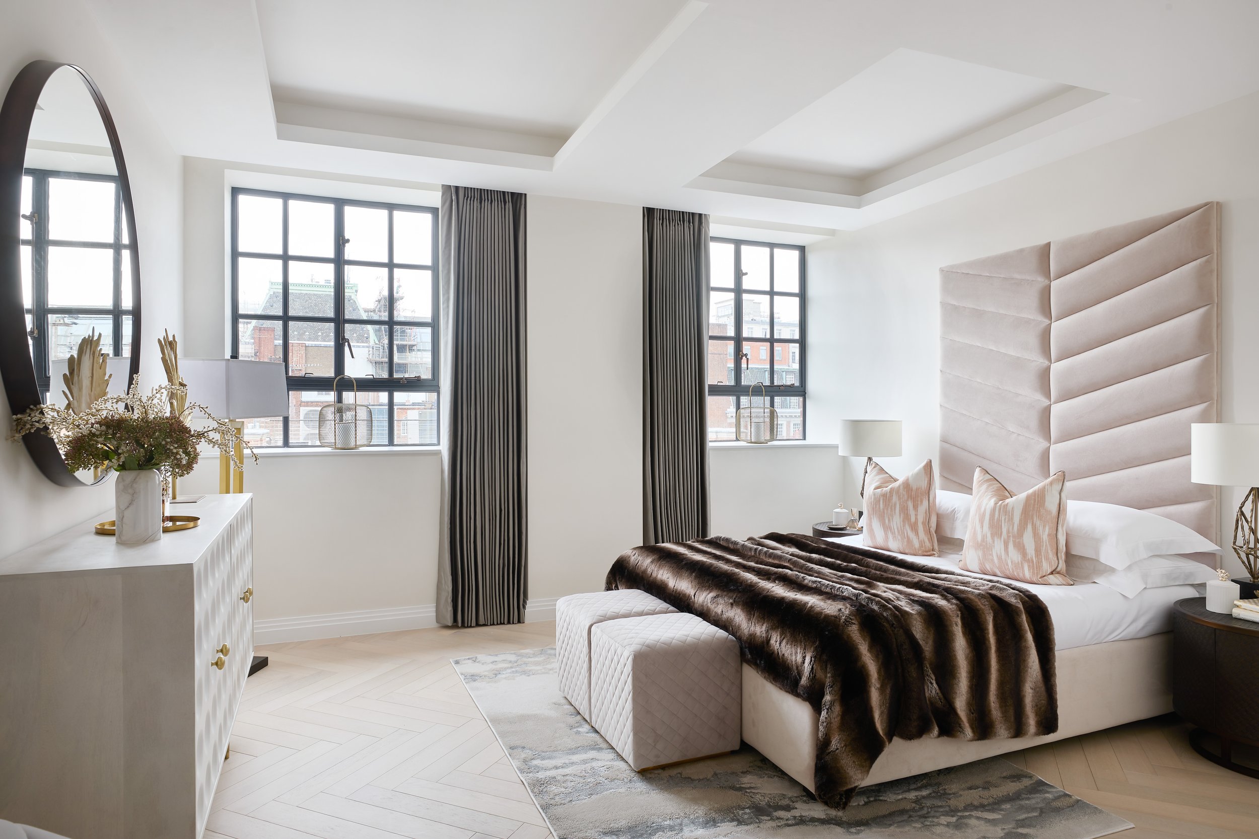 Modern bedroom with large windows, beige upholstered bed with pillows and fur throw, white dresser with vases, and gray curtains.
