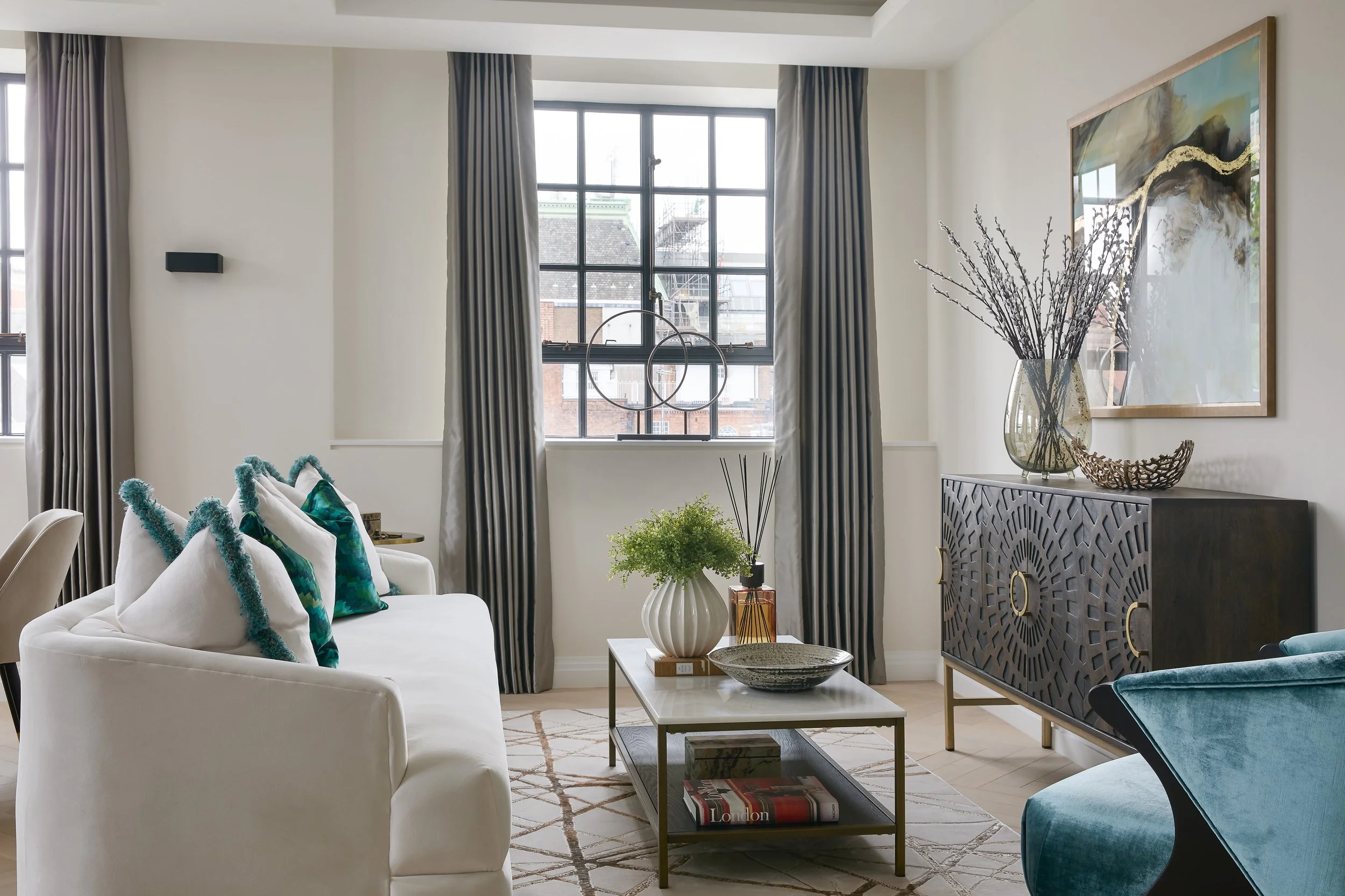 A modern living room with a white sofa, colorful pillows, a coffee table with decorative items, a sideboard with a large vase and artwork, and large windows with dark curtains.