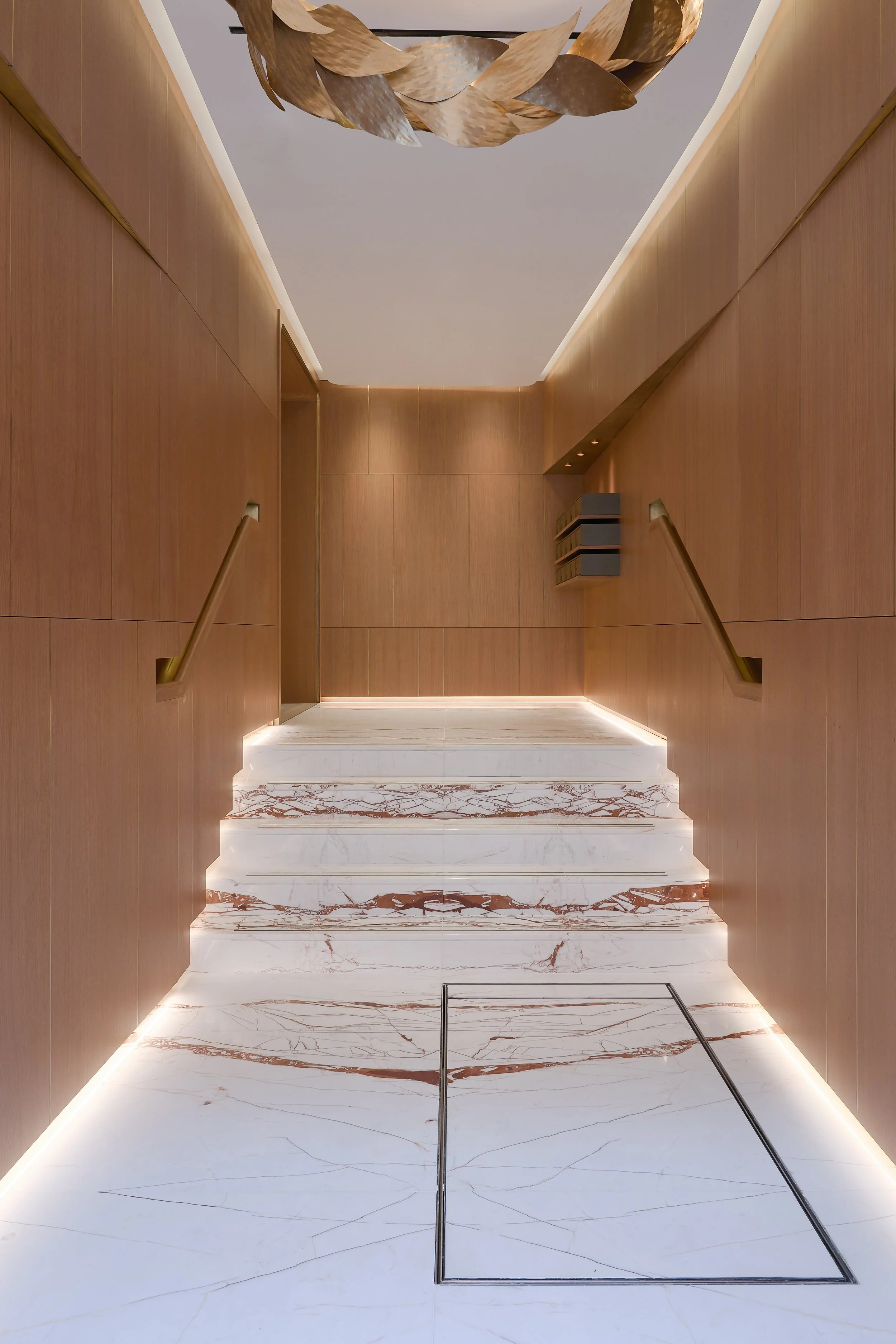 Modern interior staircase with white and brown marble steps, surrounded by wooden paneled walls, and LED lighting along the edges, leading to an upper level.