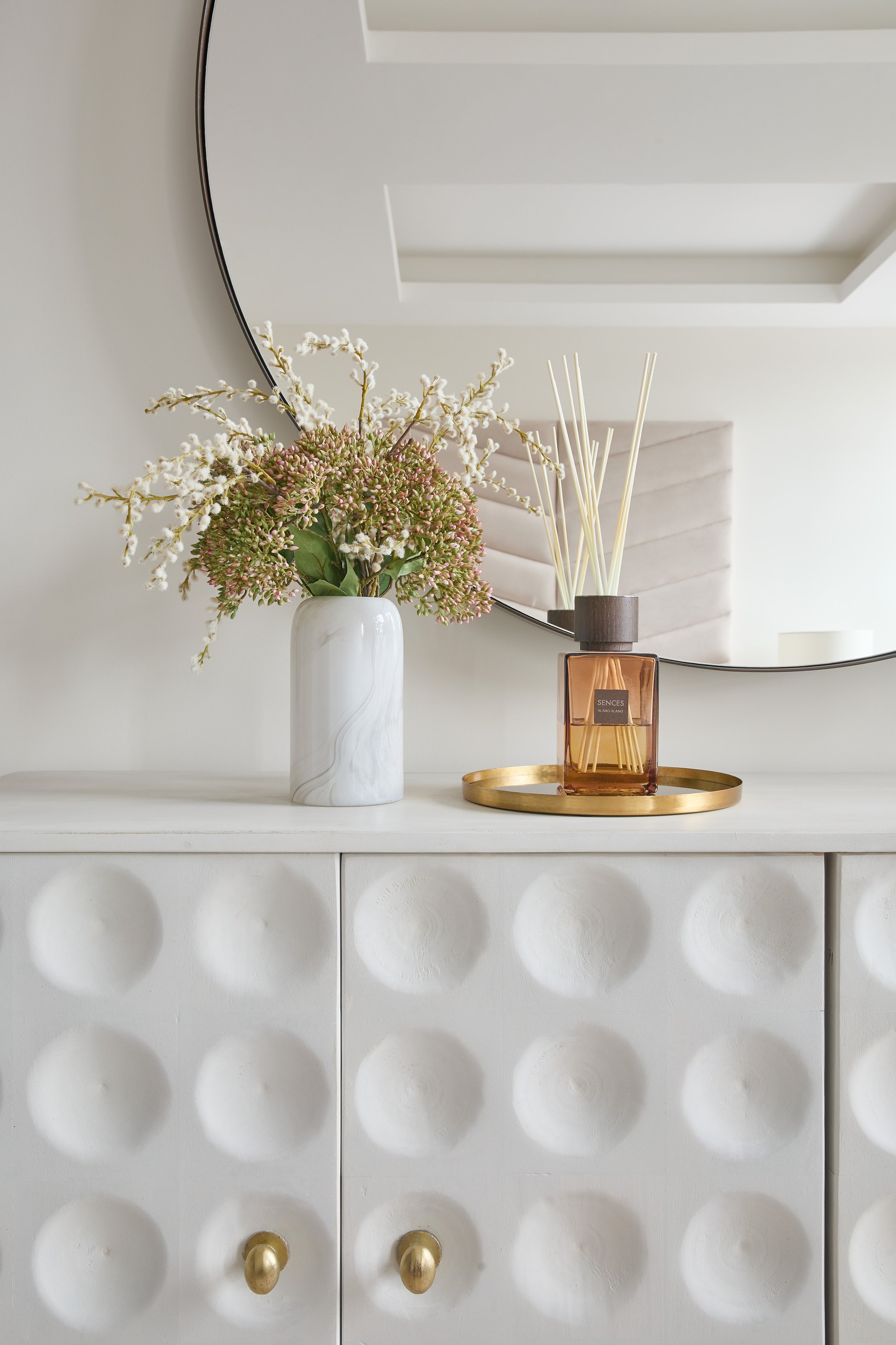 A white dresser with a textured, circular pattern on the front, topped with a marble vase containing pink and white flowers and a gold tray holding a reed diffuser with amber liquid, all reflected in a round mirror on a light wall.