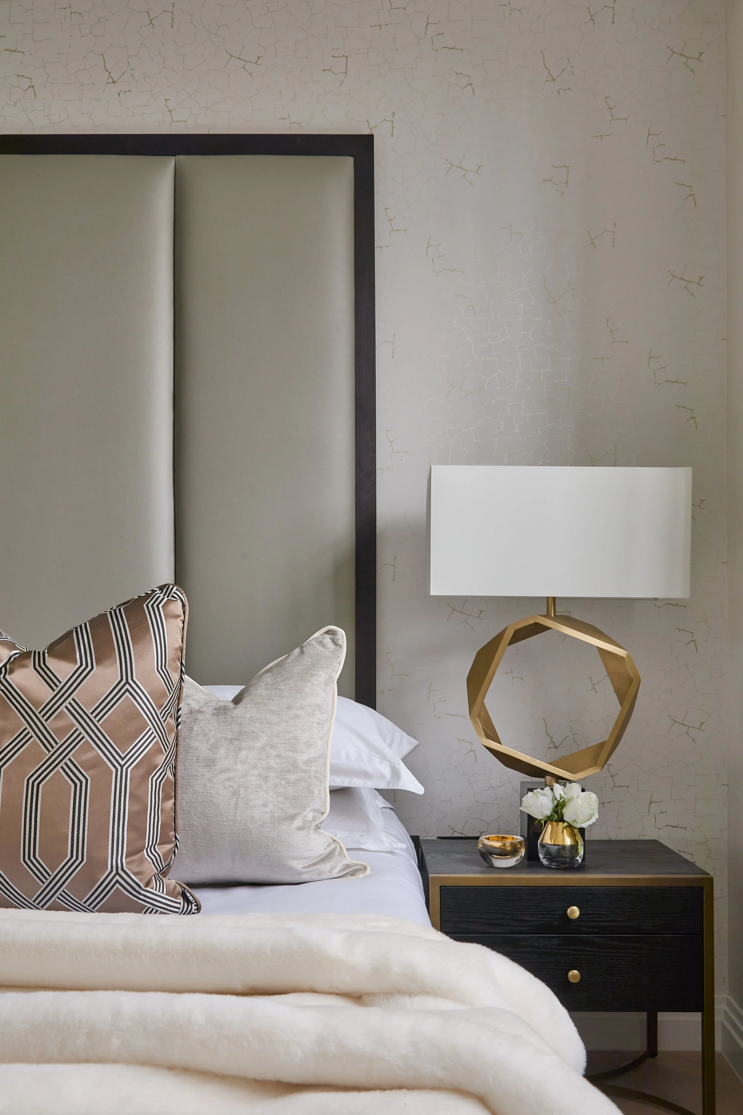 Close-up of a bedroom with a bed featuring decorative pillows, a nightstand with a vase of white roses, and a modern table lamp with a white shade and gold geometric base, against a textured wall.