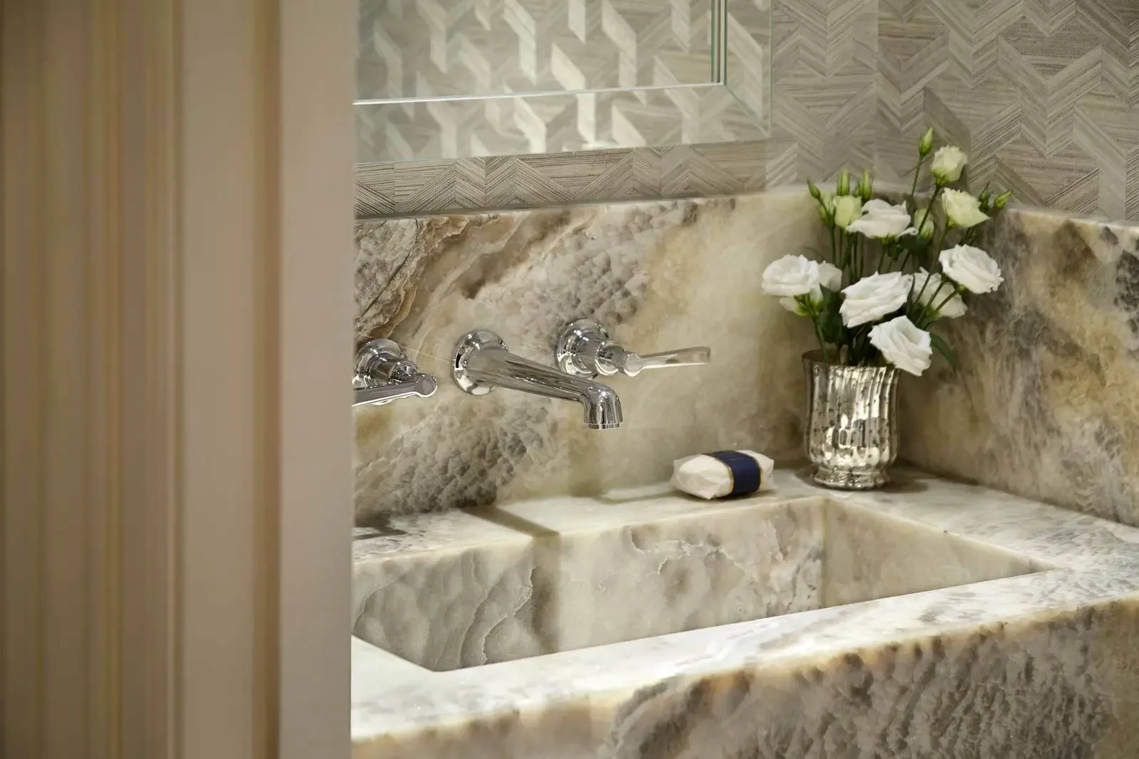 A bathroom sink with a marble countertop, a silver vase with white flowers, a bar of soap, and chrome faucets.