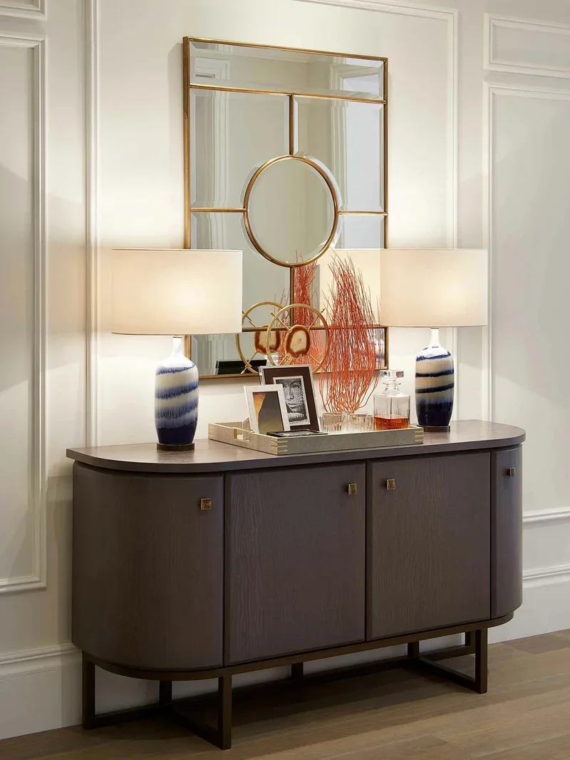 A modern console table with two table lamps, decorative picture frames, a tray, and orange decorative branches, with a rectangular mirror with gold framing hanging on a white wall above.