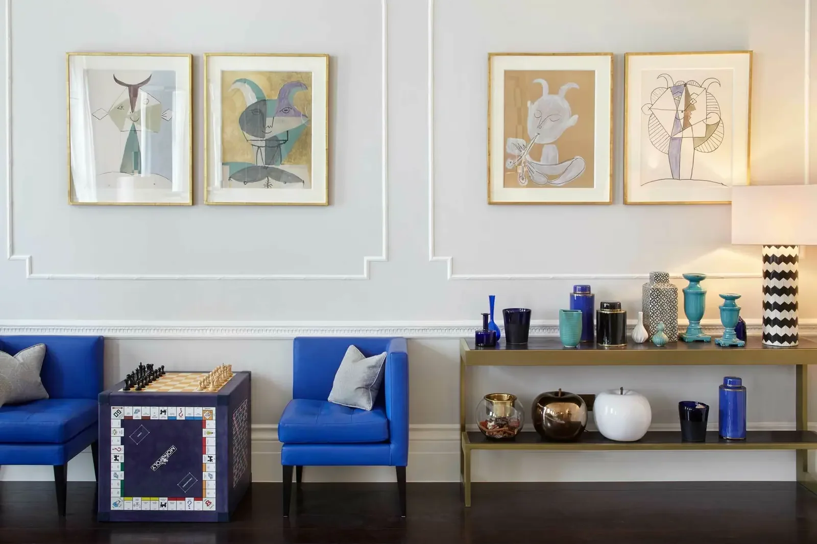 Living room with two blue chairs, a table with a chess set, and a gold and brown shelf with various blue, black, and white vases and ceramics. Four abstract art pieces hanging on the wall above the chairs.