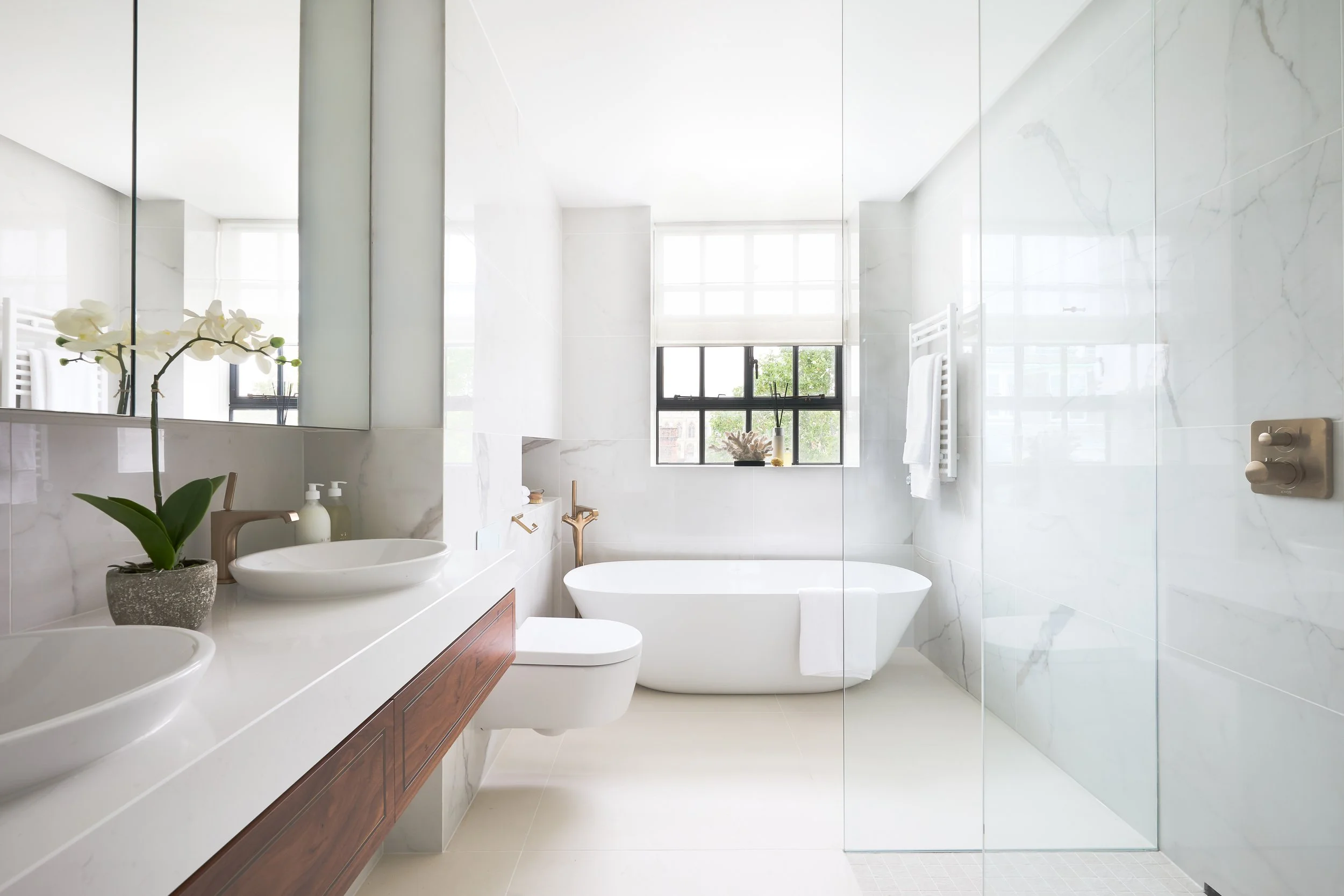 Modern white bathroom with a double sink vanity, a bathtub, a toilet, and a glass shower enclosure, decorated with a potted orchid and natural light from a large window.