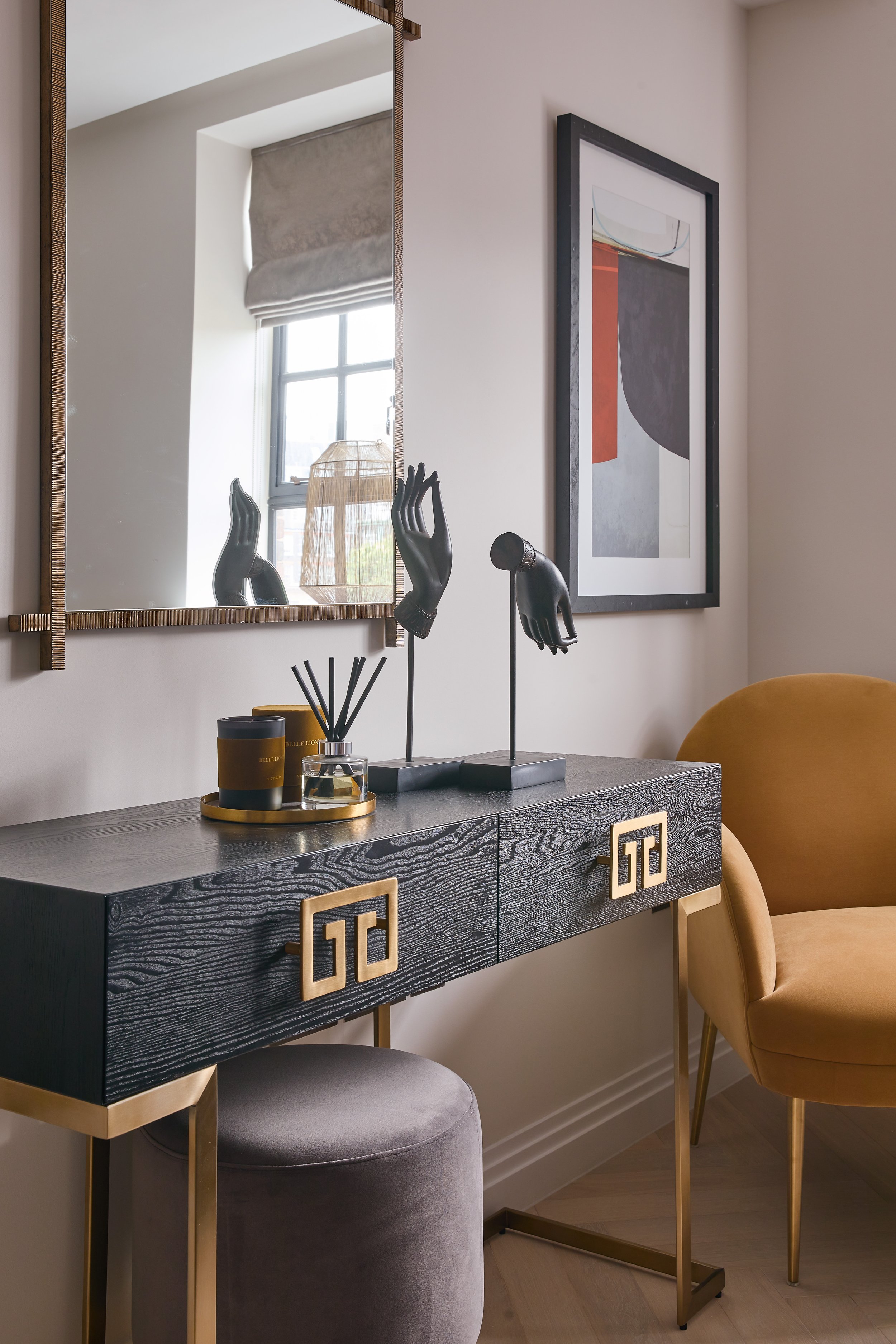 Contemporary living room with a black wooden sideboard featuring gold-tone hardware, a large wall mirror reflecting a window with a beige Roman shade and a wicker lampshade, abstract wall art, gold and black decor items, including hand sculptures and