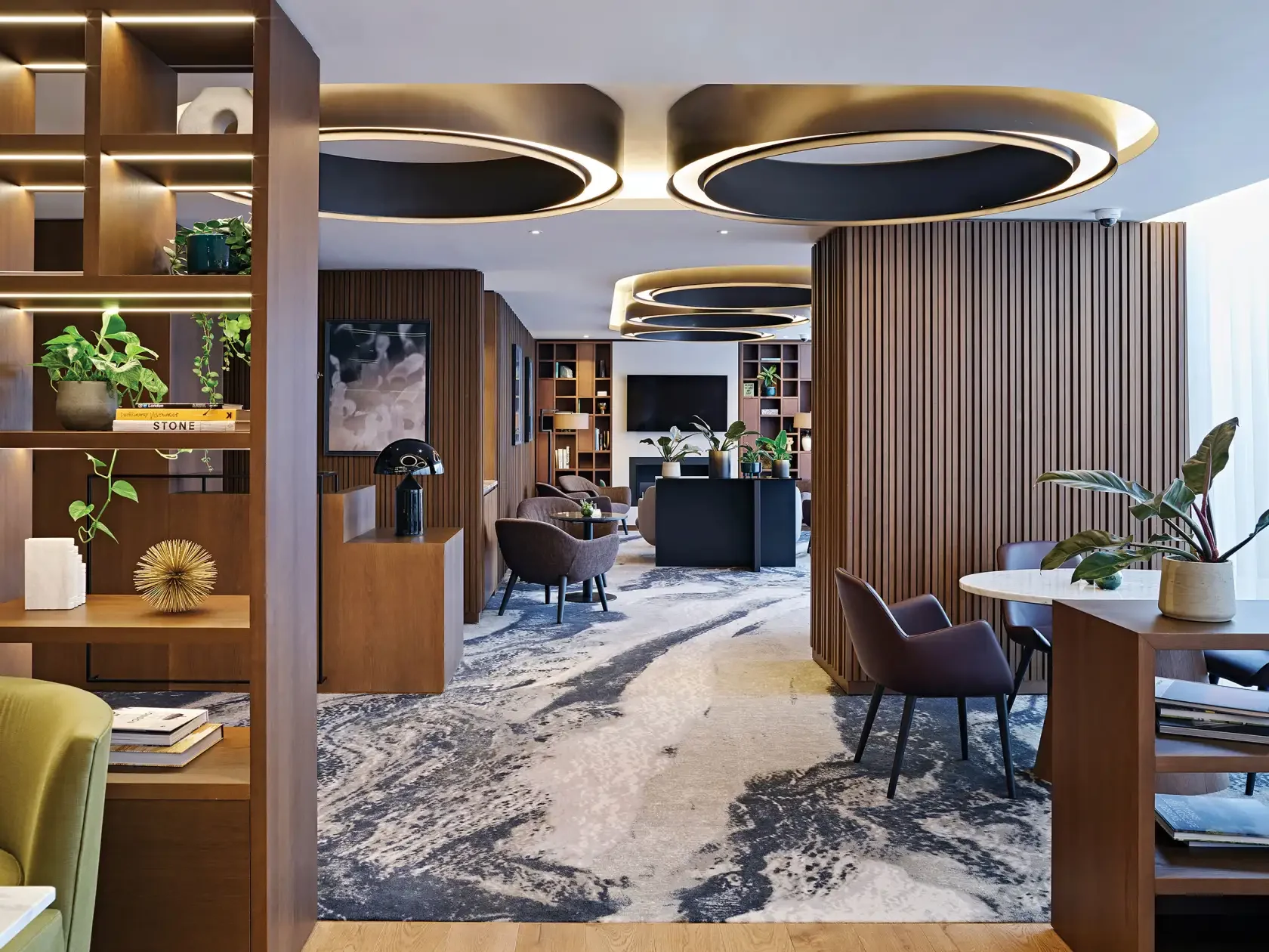 Modern hotel lobby with wooden accents, circular ceiling lights, and seating areas featuring chairs and potted plants.