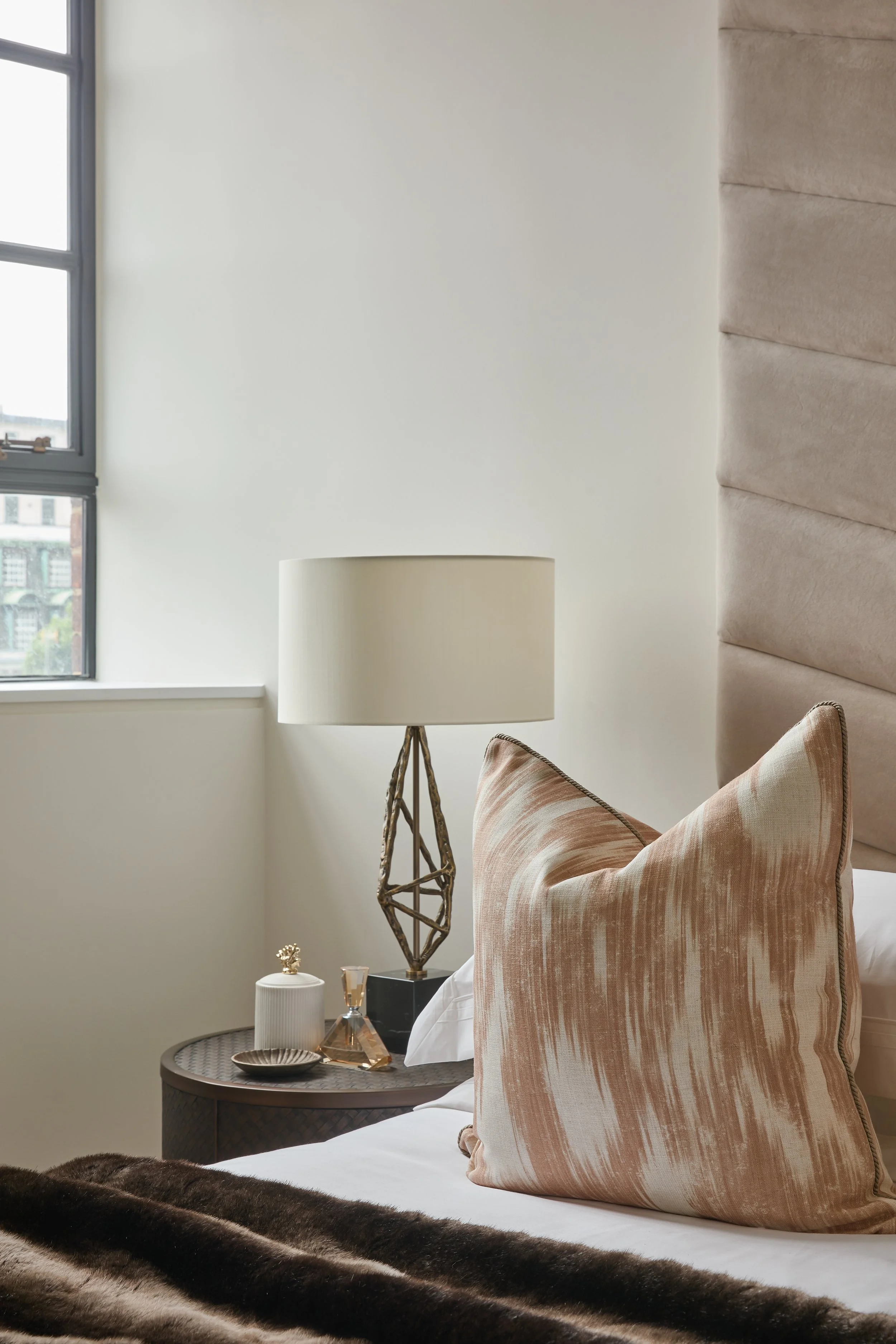 A bedside table with a decorative lamp, small decor items, and a cushion on a bed in a bedroom with a large window and soft lighting.