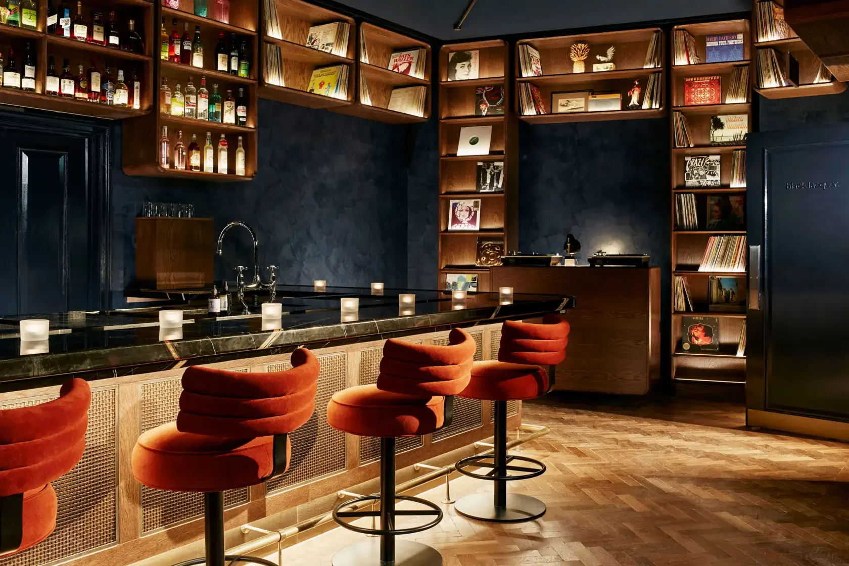 A cozy bar with red velvet bar stools, a black marble counter, and dark blue walls decorated with shelves of vinyl records and books.