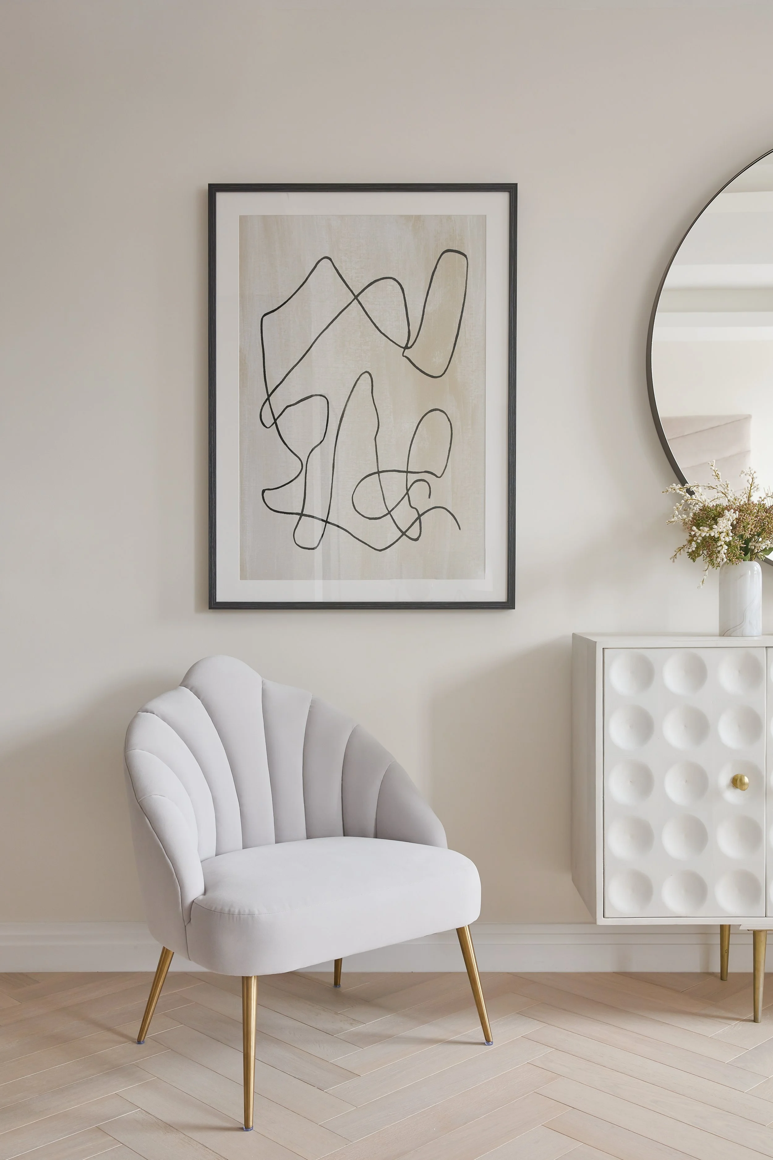 A white upholstered armchair with gold legs sits on a light wood floor next to a white cabinet with a circular pattern and a gold knob. Above, a framed abstract line drawing hangs on a beige wall, and a round mirror partially reflects the room. A whi