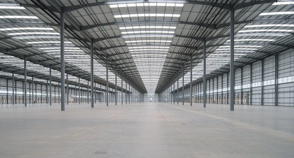 Empty, spacious industrial warehouse with metal roof, glass panels, and supporting columns.