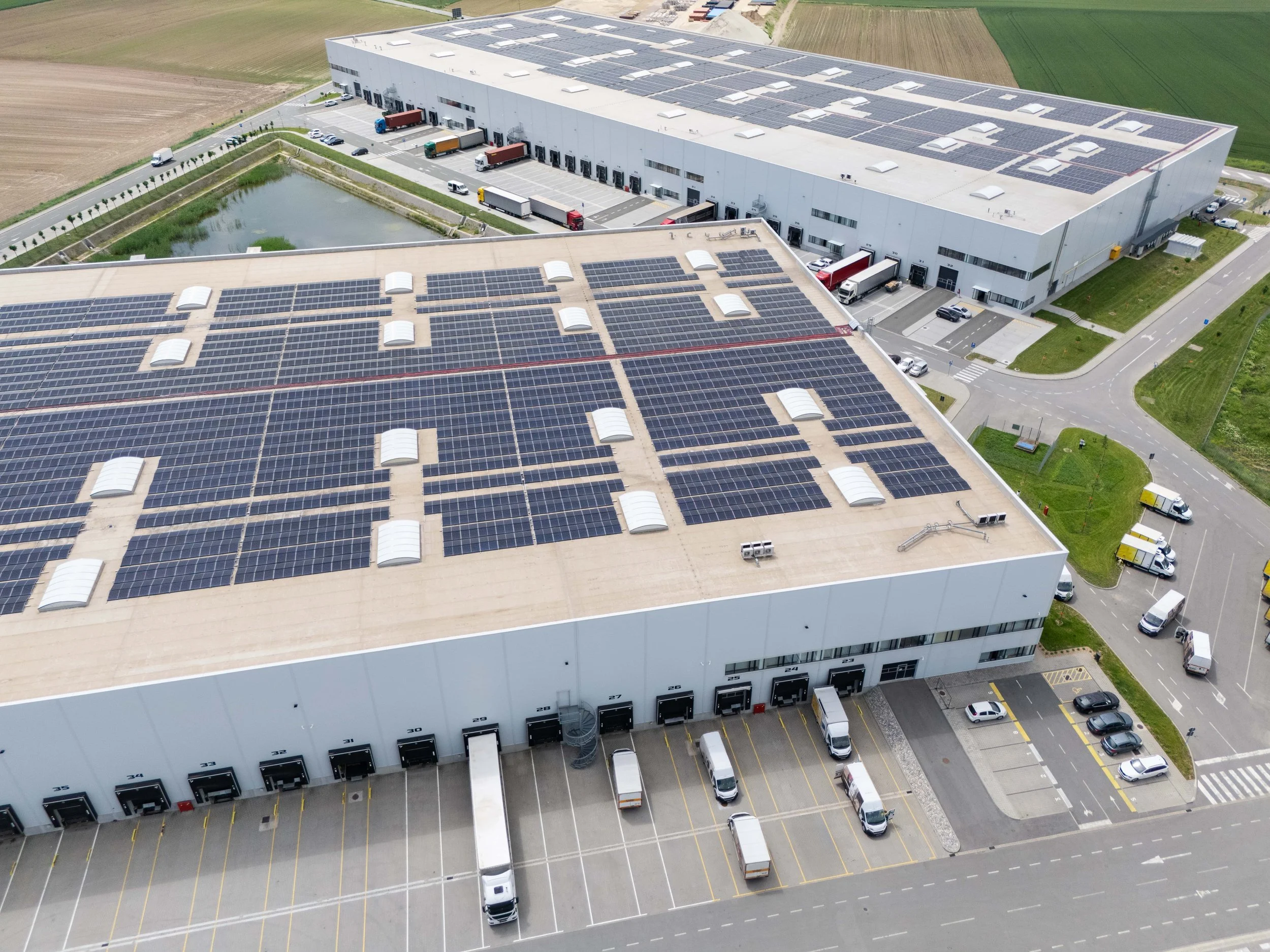 Aerial view of a commercial warehouse building with solar panels on the roof, loading docks, parked trucks and cars, and surrounding roads and fields.