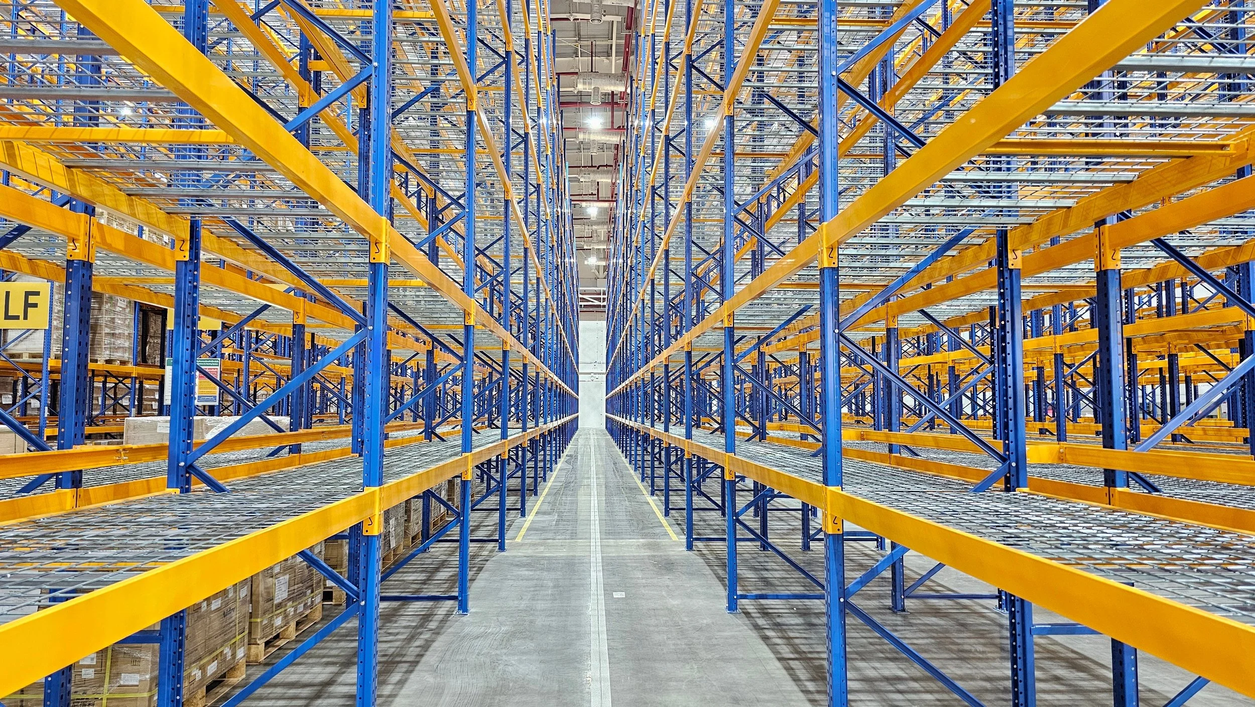 Empty warehouse with tall blue and yellow metal shelves and a concrete aisle in the center.