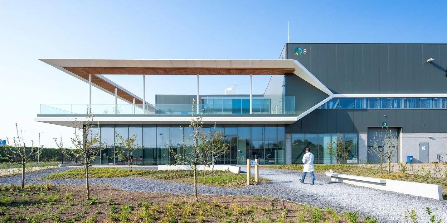 Modern building with large glass windows, a man walking towards the entrance, and trees in the landscaped foreground.