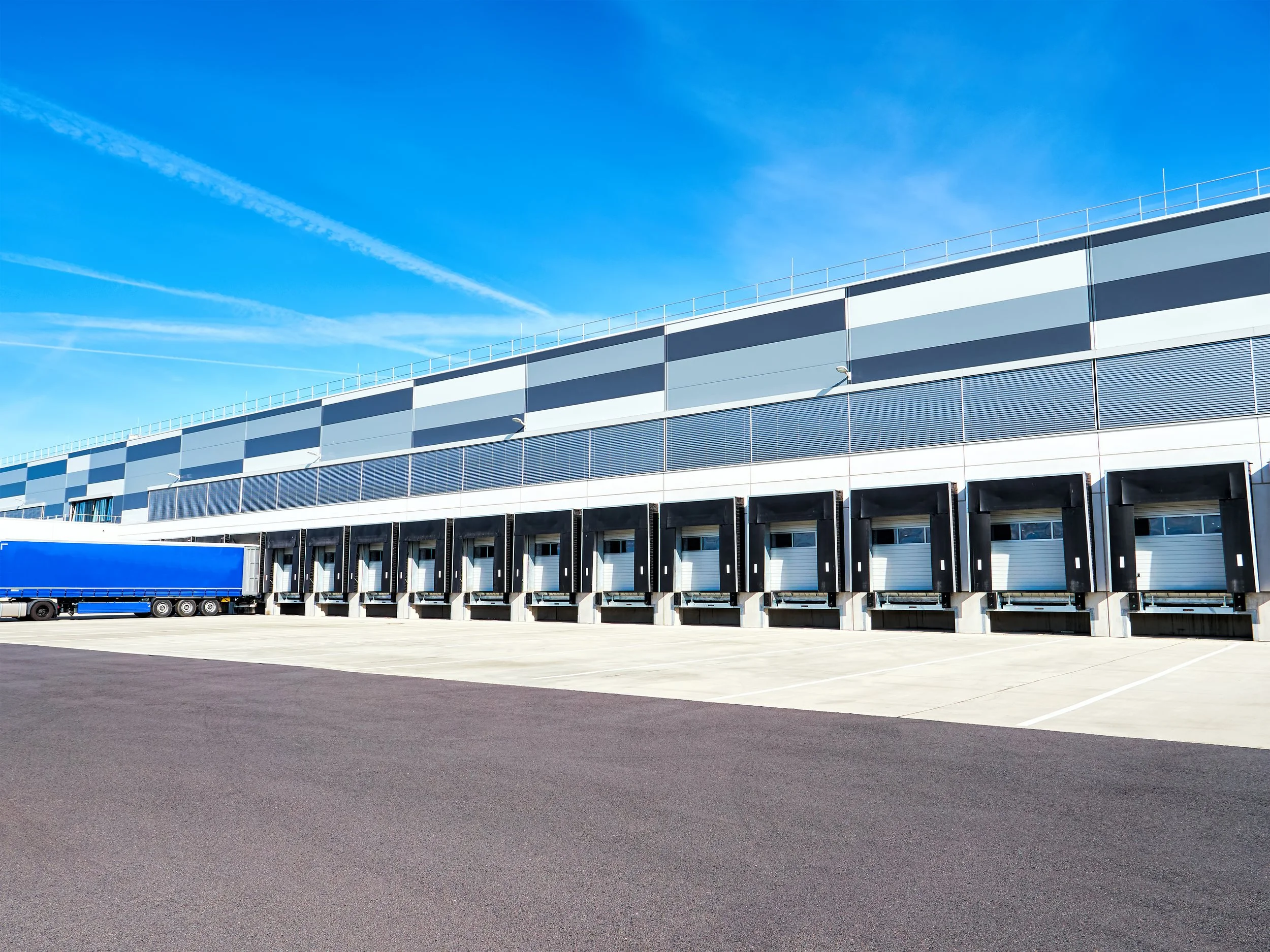 A large modern warehouse or distribution center with multiple loading docks and a blue truck parked in front under a clear blue sky.