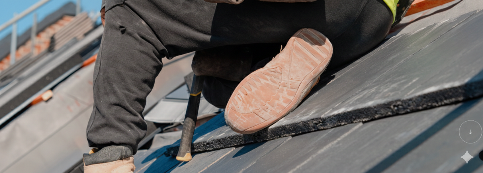 Close-up of a person in work clothes installing or repairing a roof, kneeling on shingles with a hammer and wearing a work glove.
