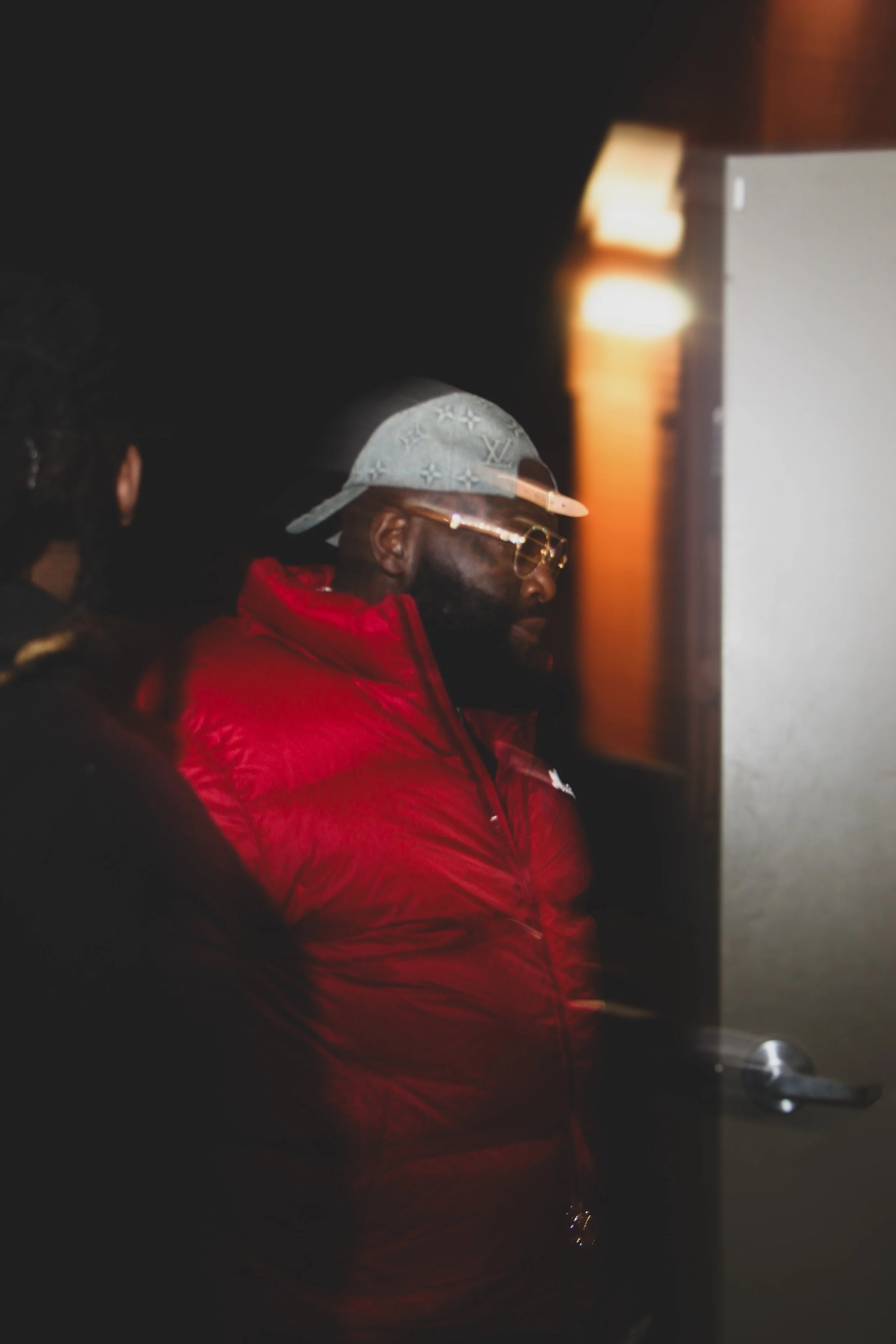 A man wearing sunglasses, a light gray cap, and a red jacket is standing indoors near a door, looking at a monitor screen with a woman partially visible next to him.