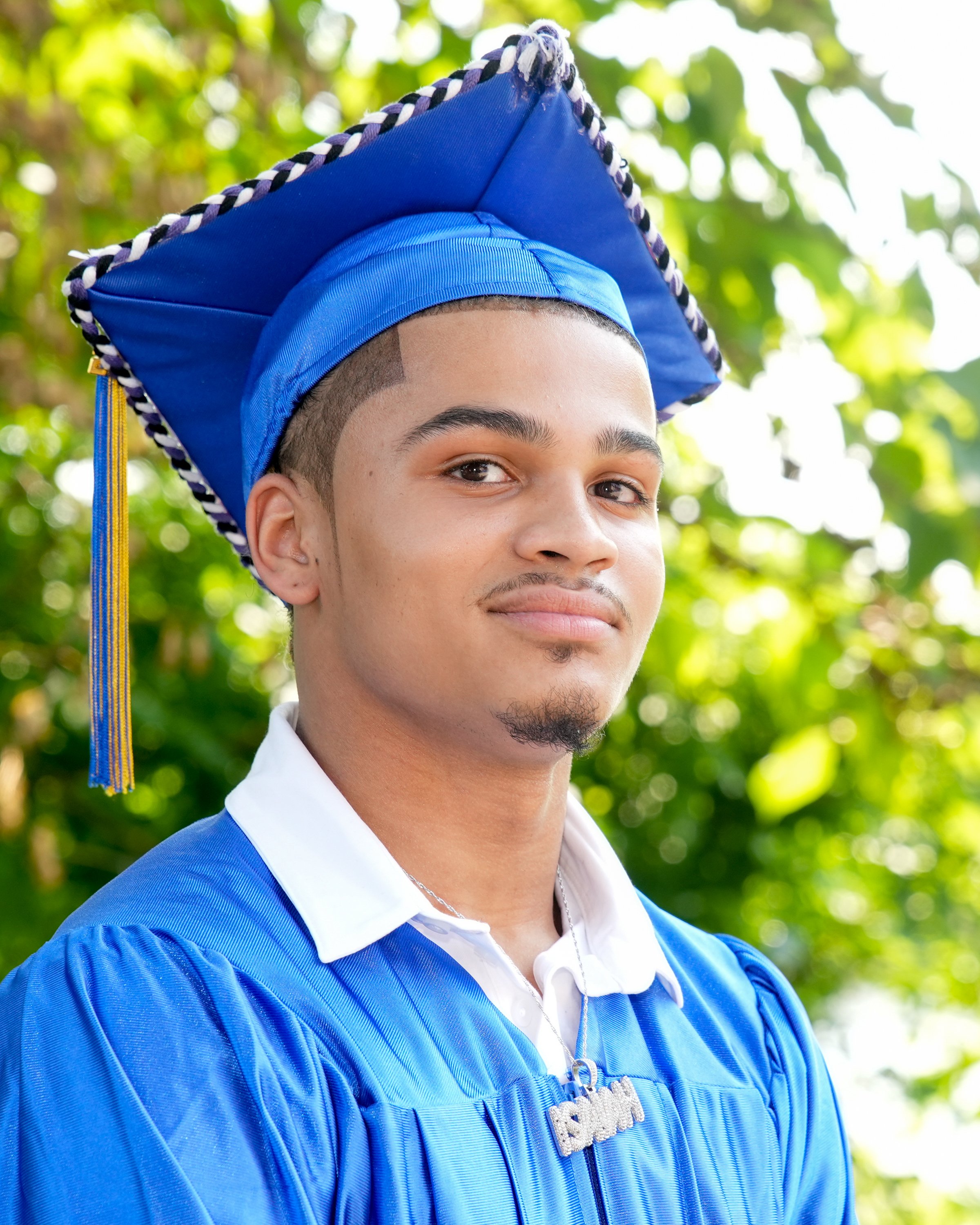 Young man in a blue graduation gown and cap, outdoors with green leafy background.