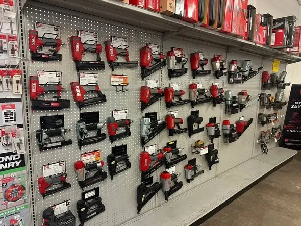 A store display of various cordless nail guns mounted on a white pegboard, with shelves above and below.