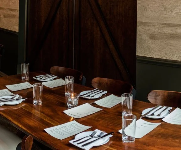 A wooden dining table set for a meal, with papers, drinking glasses, and cutlery arranged at each place setting, in a room with dark wood and wood-paneled walls.