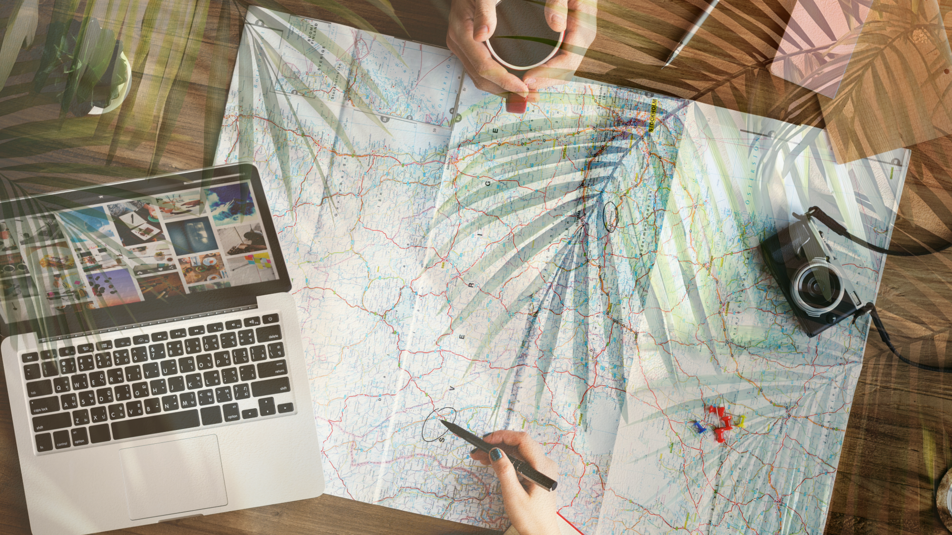 Top-down view of a workspace with a map, laptop, camera, and a hand holding a coffee cup.