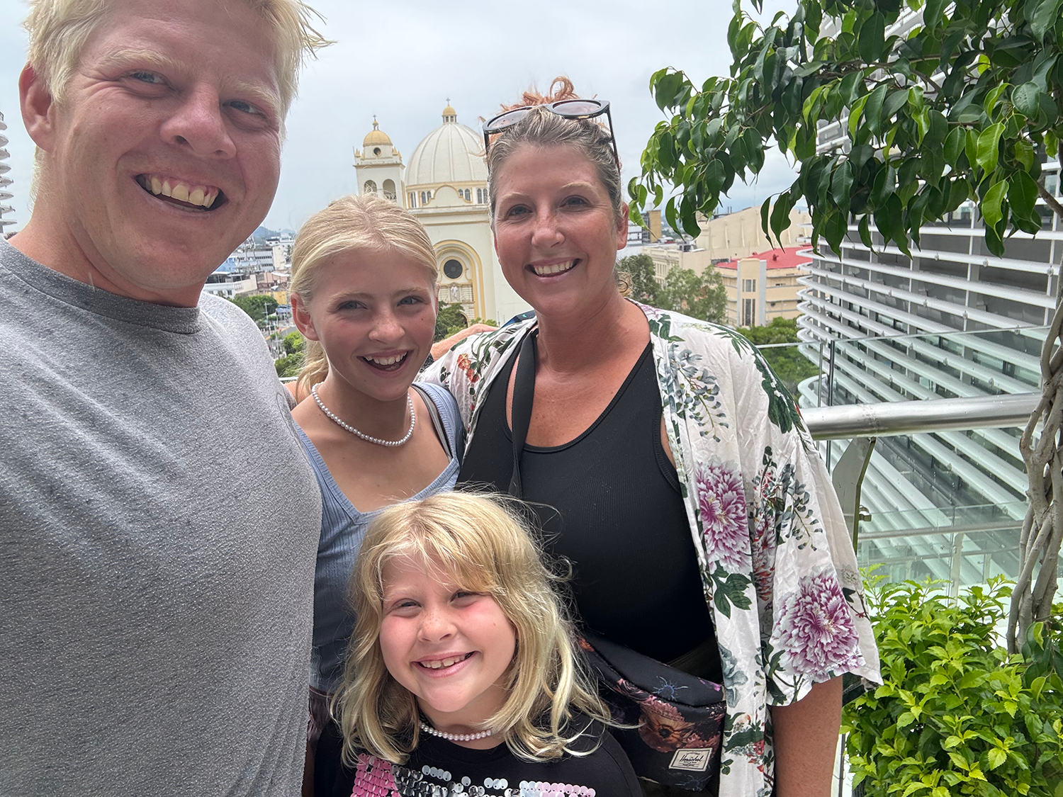 Gulyas family of four taking a selfie on a balcony with a cityscape and a church with domes in the background, greenery, and modern buildings.